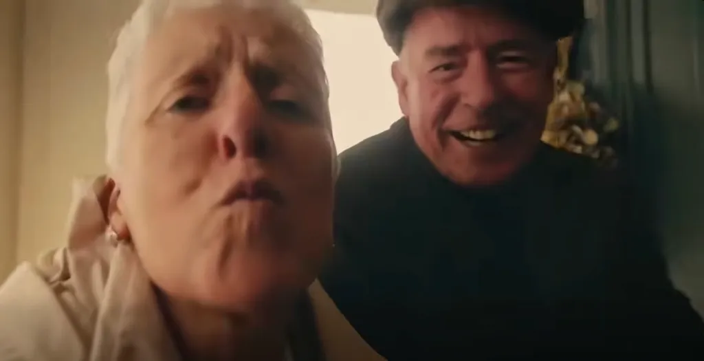 An older woman with short white hair leans in closely with her lips pursed, while an older man in a cap stands behind her, smiling.