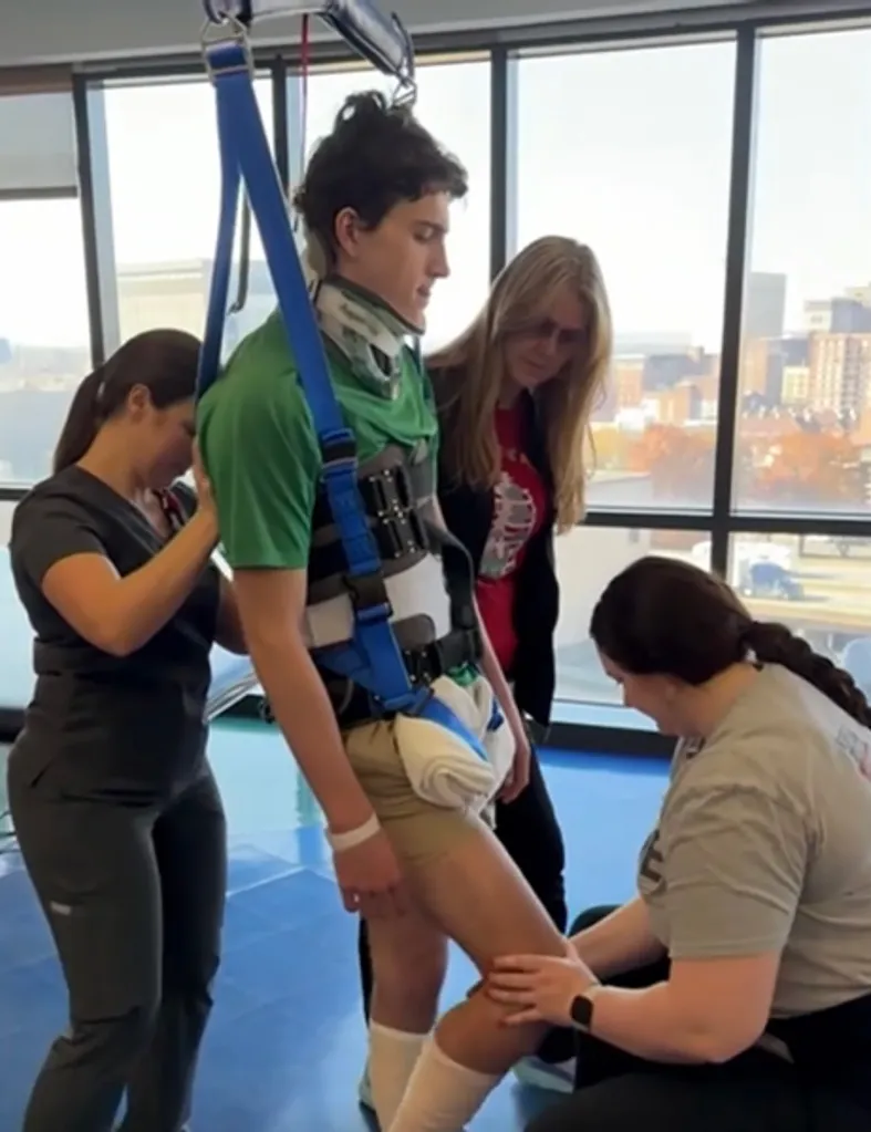 Wyatt Anderson, 17, receiving physical therapy with a harness and neck brace, supported by medical staff and another person.