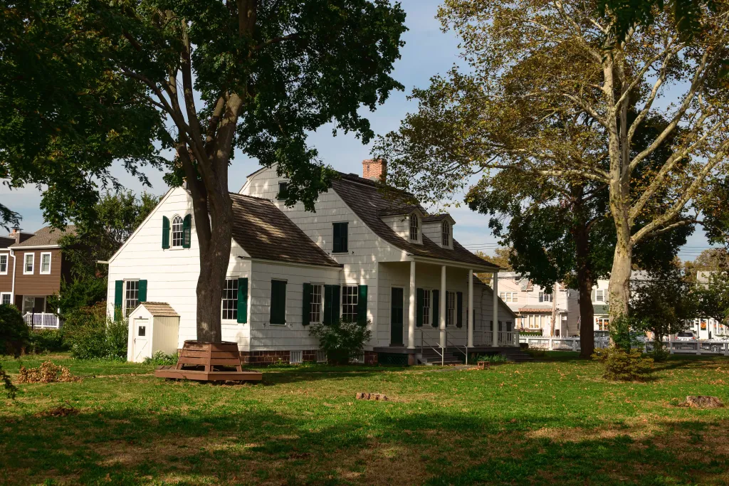 The historic Hendrick I. Lott House, a white Dutch-American farmhouse with green shutters, stands surrounded by trees and a grassy lawn in the Marine Park neighborhood of Brooklyn.