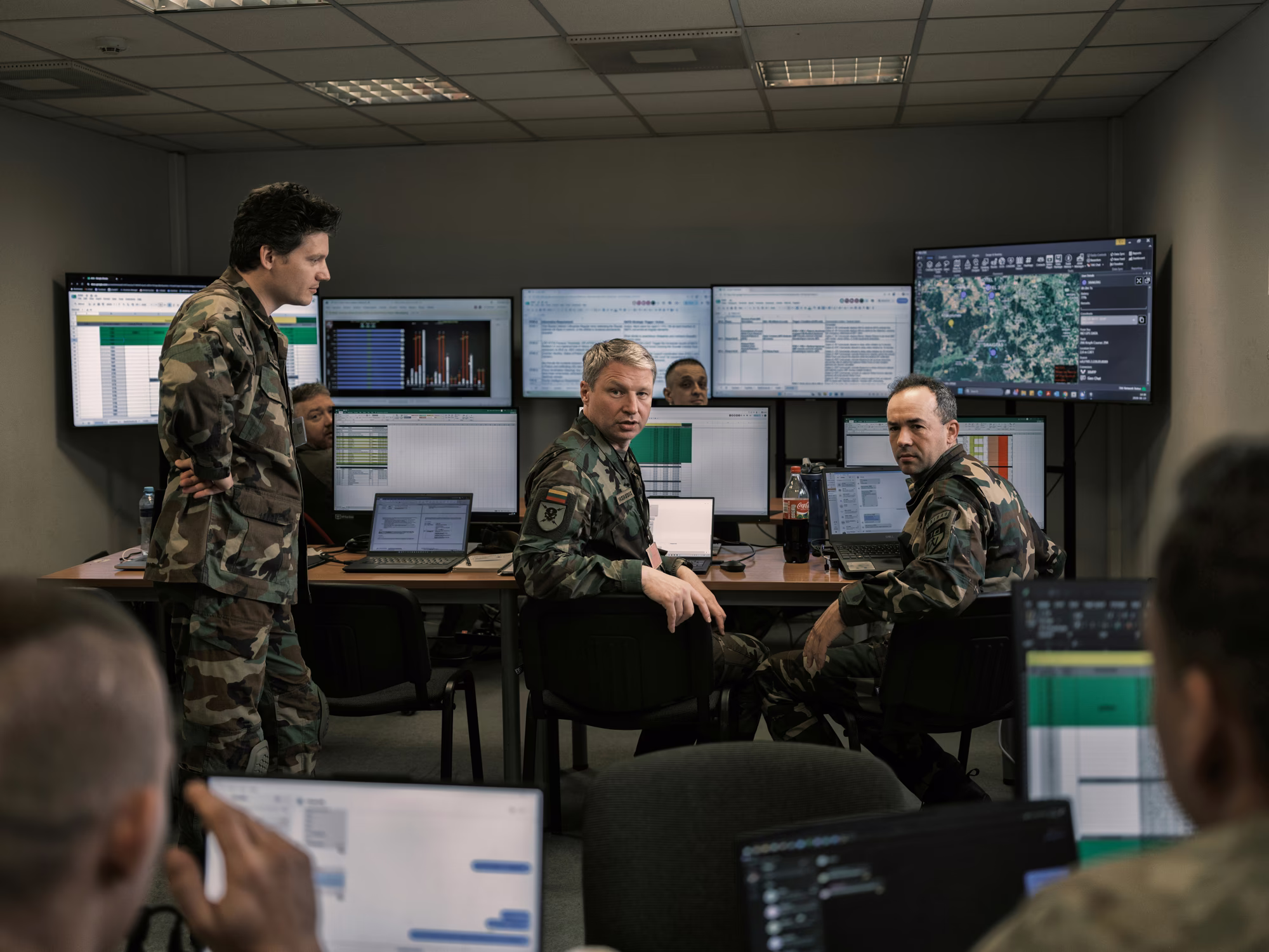 photo of group of uniformed people sitting at conference table with numerous monitors behind them, talking with additional militia members holding laptops in foreground