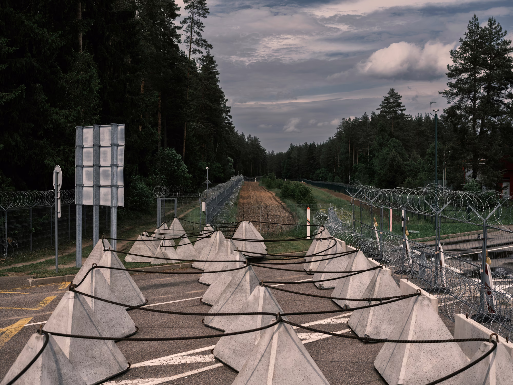 photo of road at border area crossing with rows of large pyramidal concrete blocks linked together, layers of fences with razor wire surrounding cleared dirt areas, and forest in background