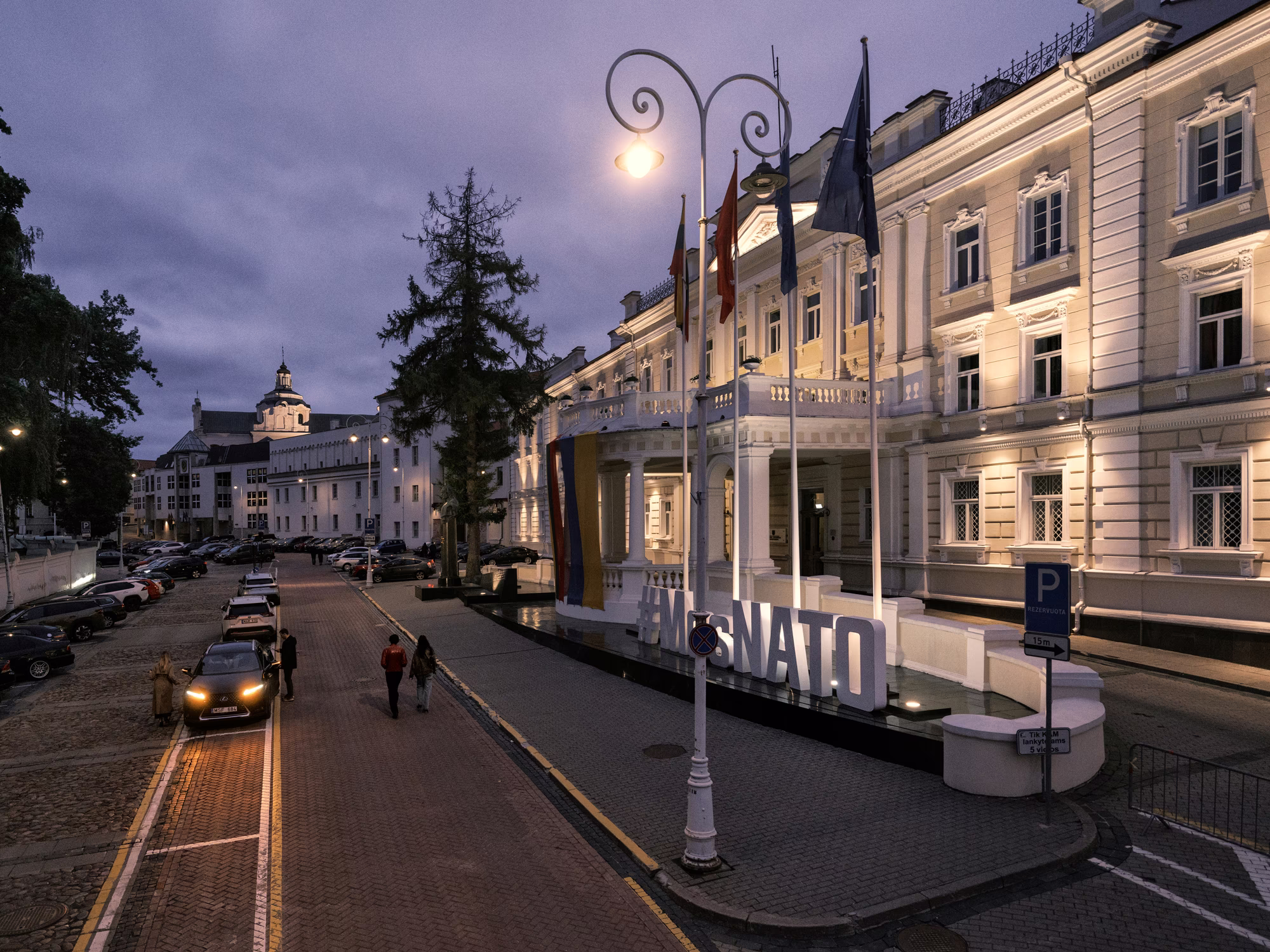 photo at twilight of large ornate building on right with flags and large #MesNATO sign, with parked cars and people walking along brick street alongside it