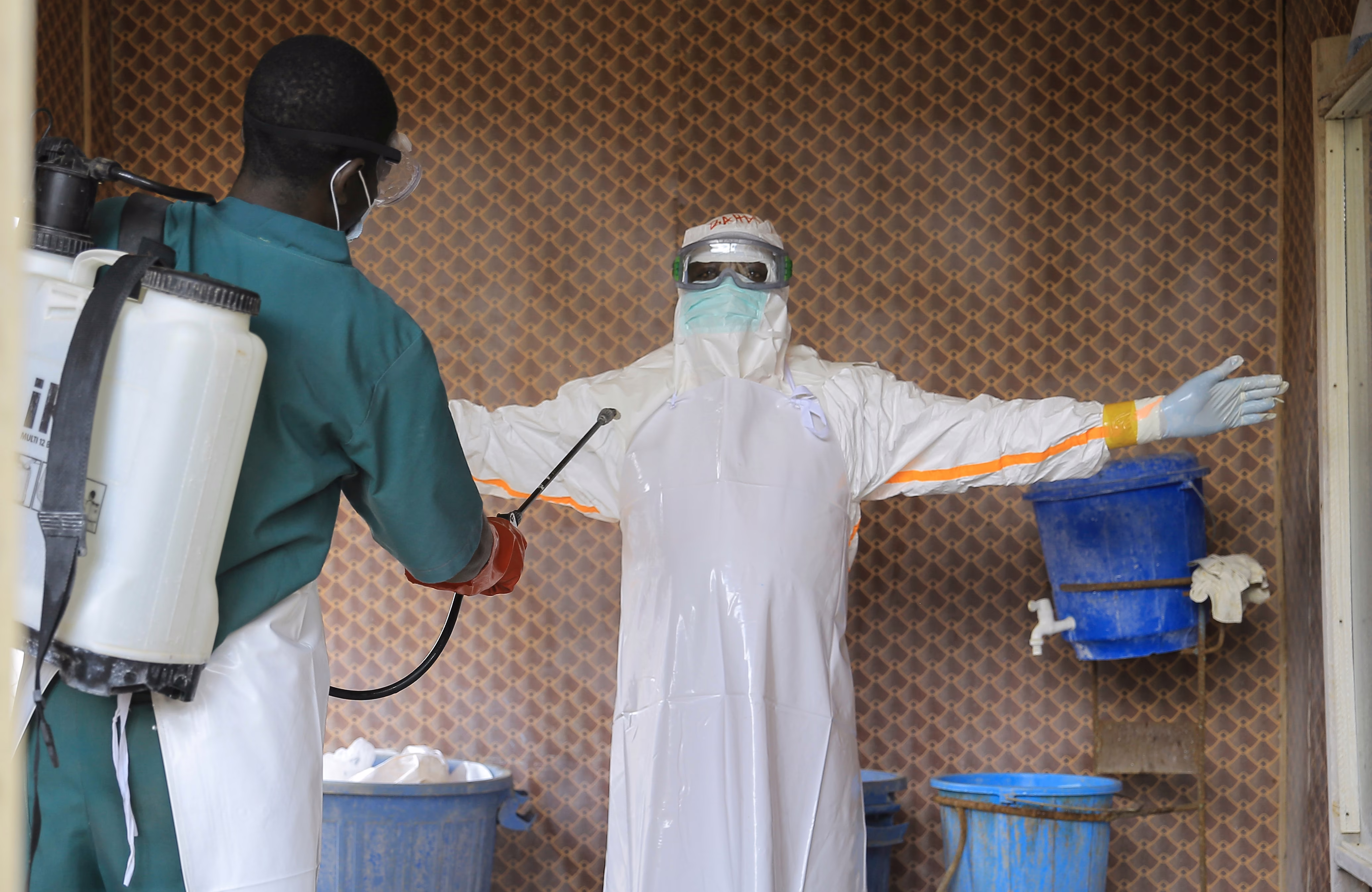 A healthcare worker in protective clothing is sprayed with disinfectant by a colleague.
