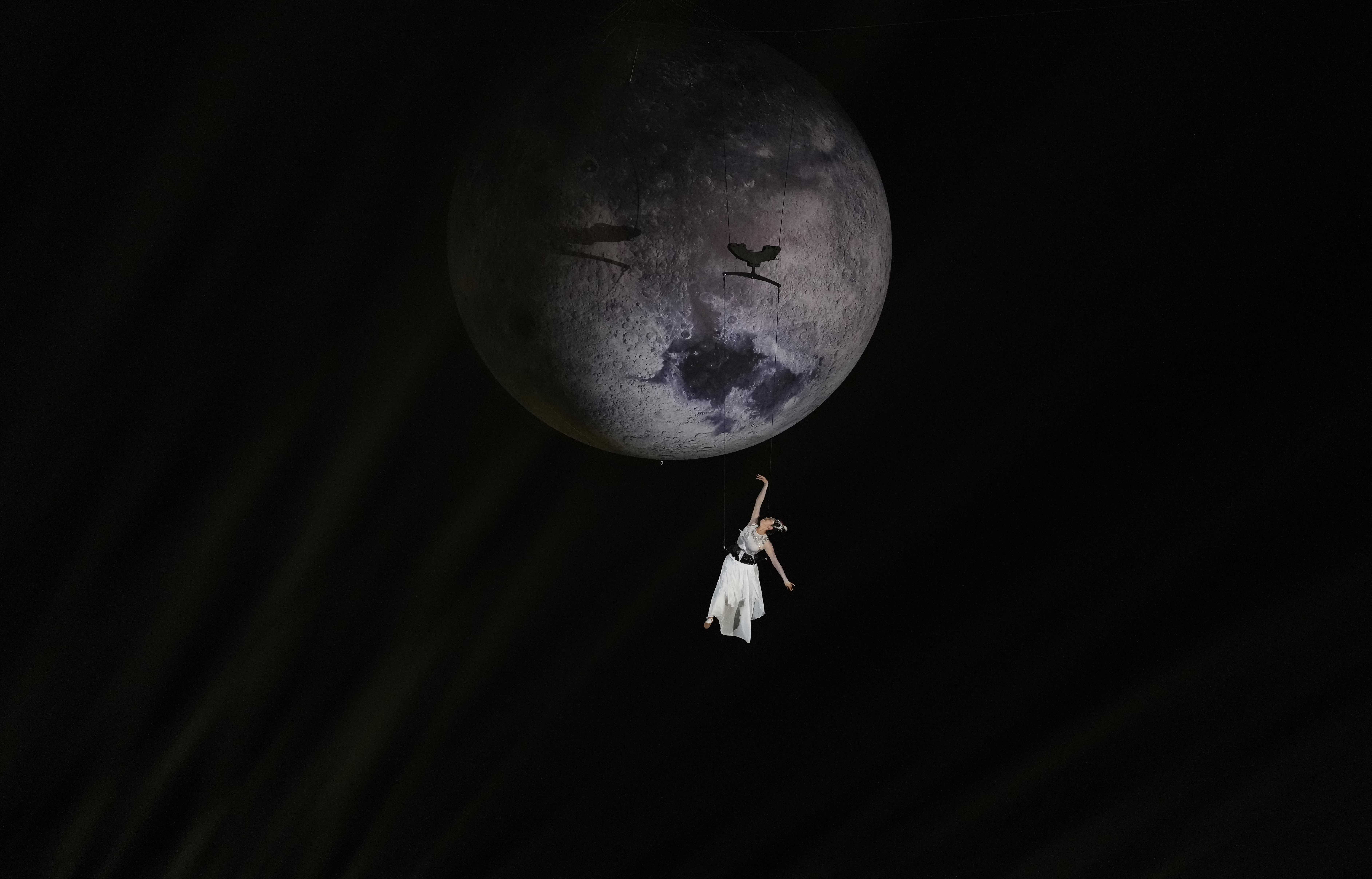A performer hangs from wires, appearing to fly beneath the moon.