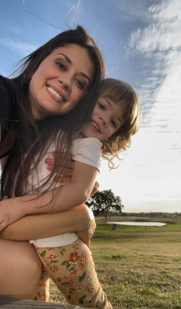 Cibelle Savella holding her daughter Vivian Savella outdoors against a bright sky.