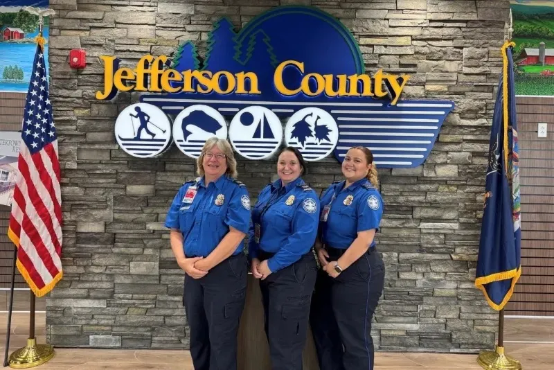 TSA officers Yessenia Alvarez, Melissa Graffam, and Debra Alcock pose for a photo in front of a Jefferson County sign flanked by American and state flags.