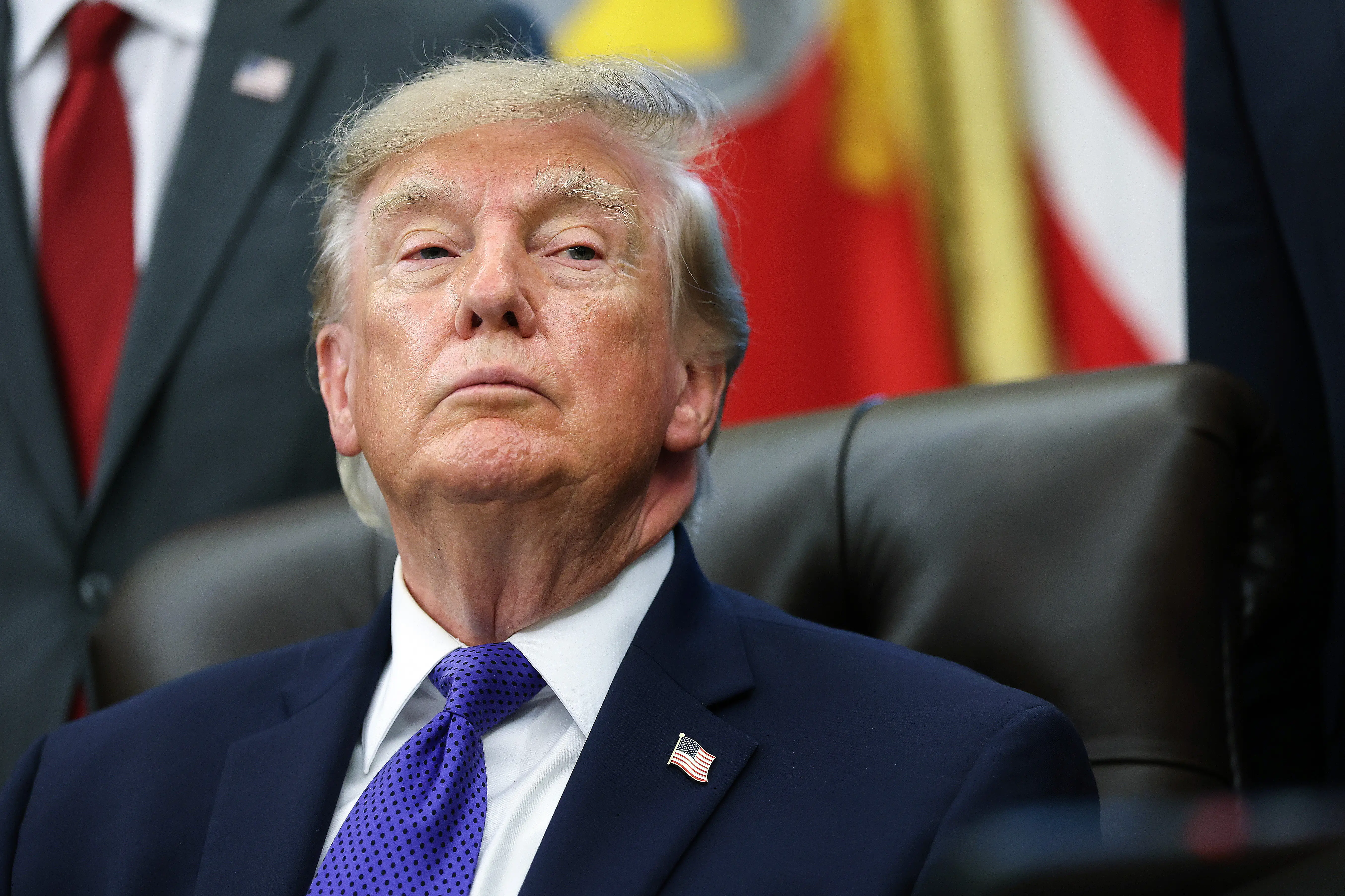 Donald Trump sits in a leather chair with flags and suited people visible behind him.