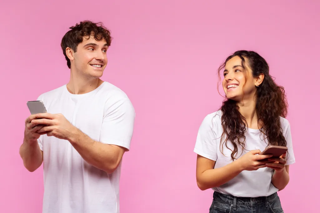 A smiling man and woman holding smartphones and looking at each other against a pink background.
