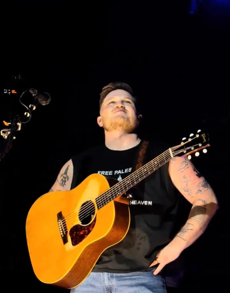 Zach Bryan holding an acoustic guitar on stage.