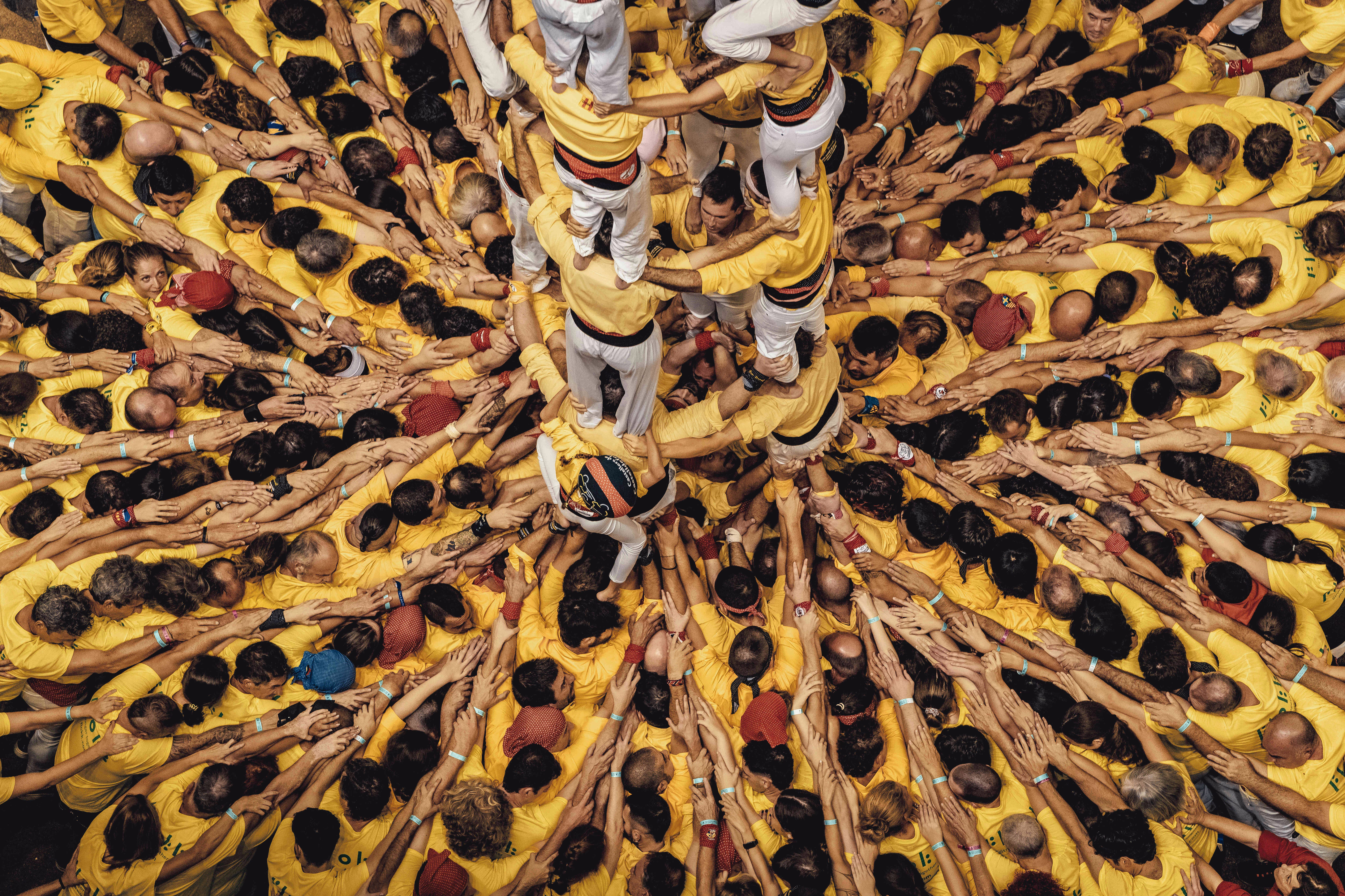 An overhead view of a large crowd of people wearing yellow, pusing inwards, arms linked over each other's shoulders, supporting a human tower in the center