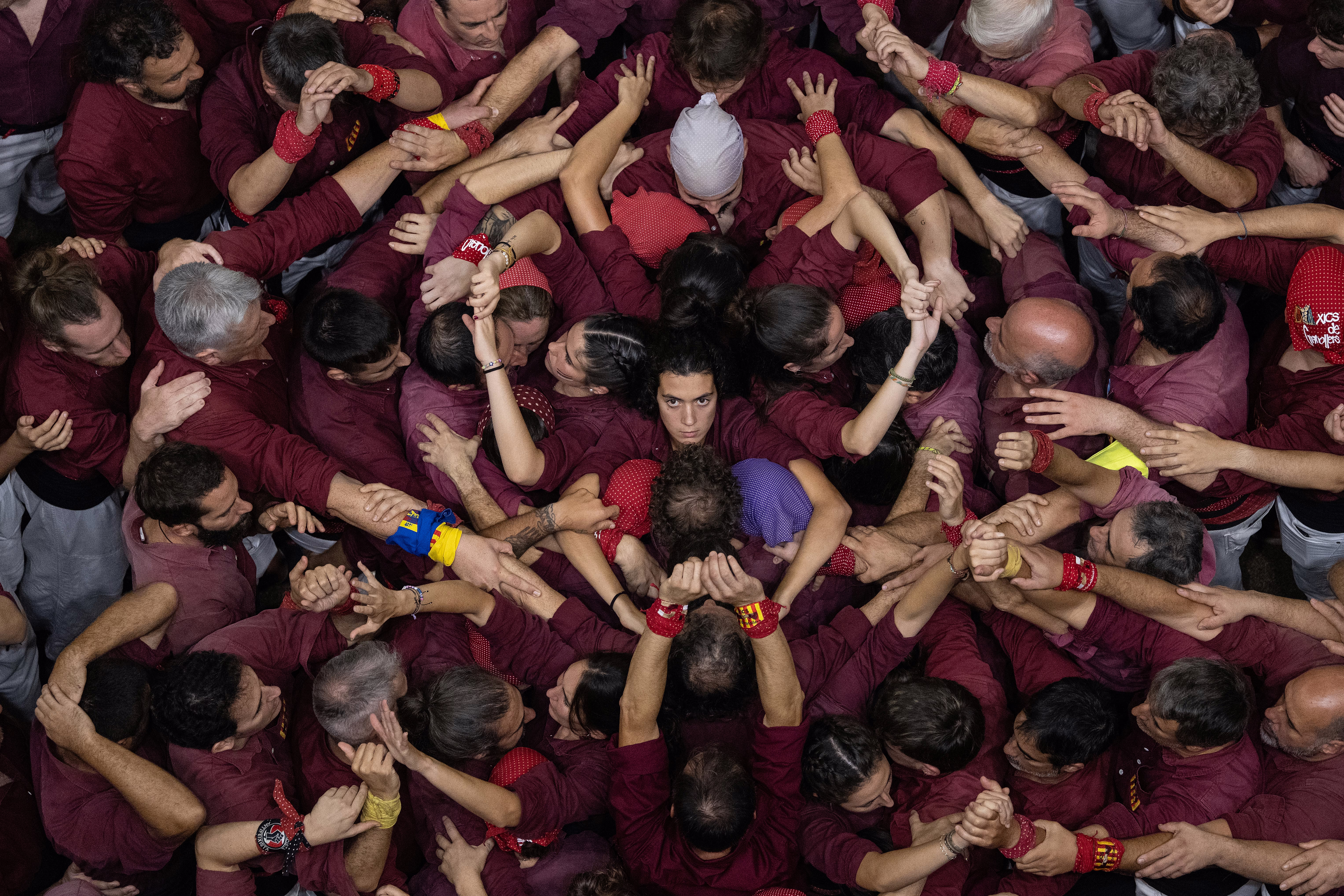 An overhead view of a group of people wearing purple shirts, preparing to build a human tower. One person at center looks straight up toward the camera.