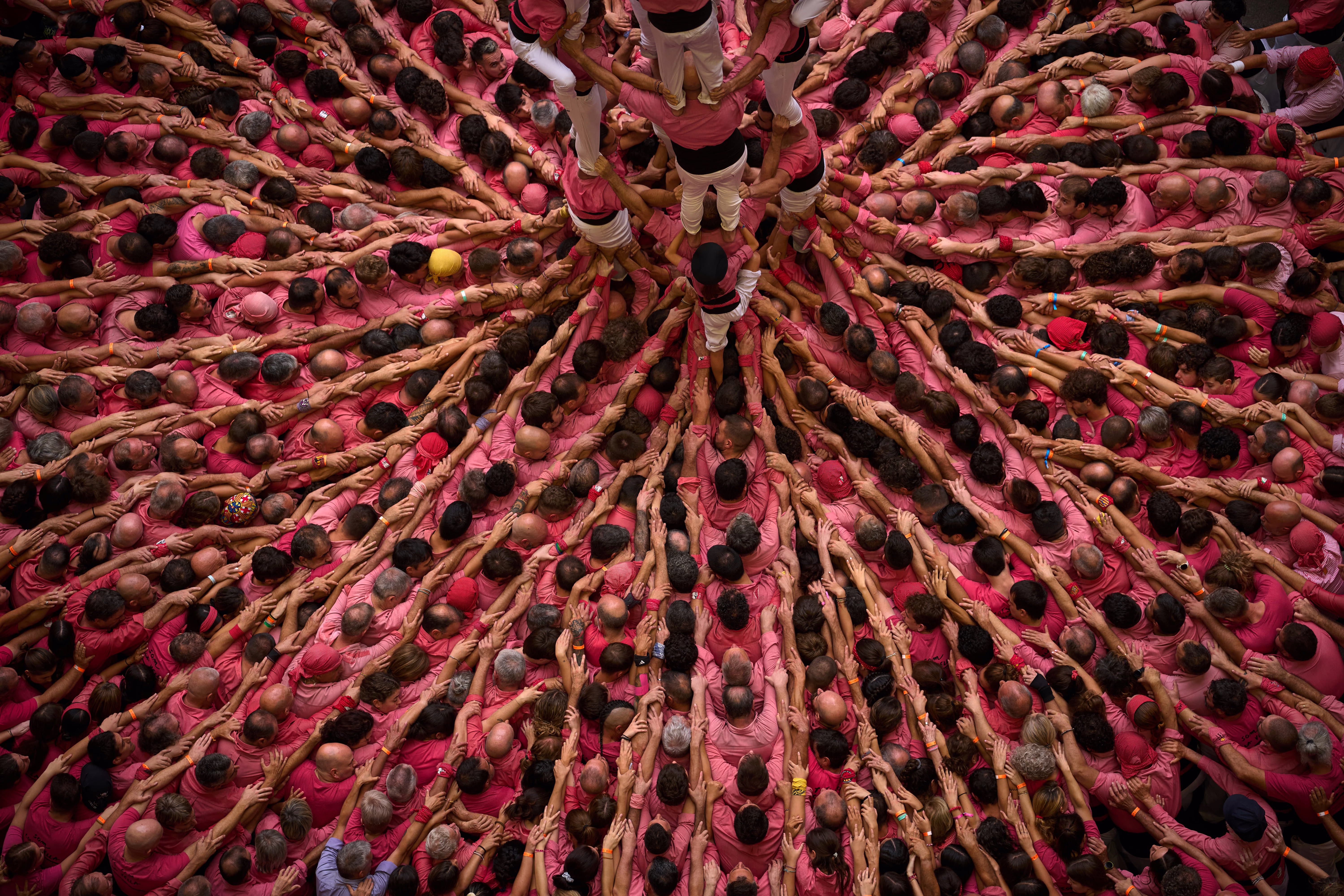An overhead view of a large crowd forming a human tower. Many people lined up, pointing toward the center of the group, with arms linked over each other's shoulders.