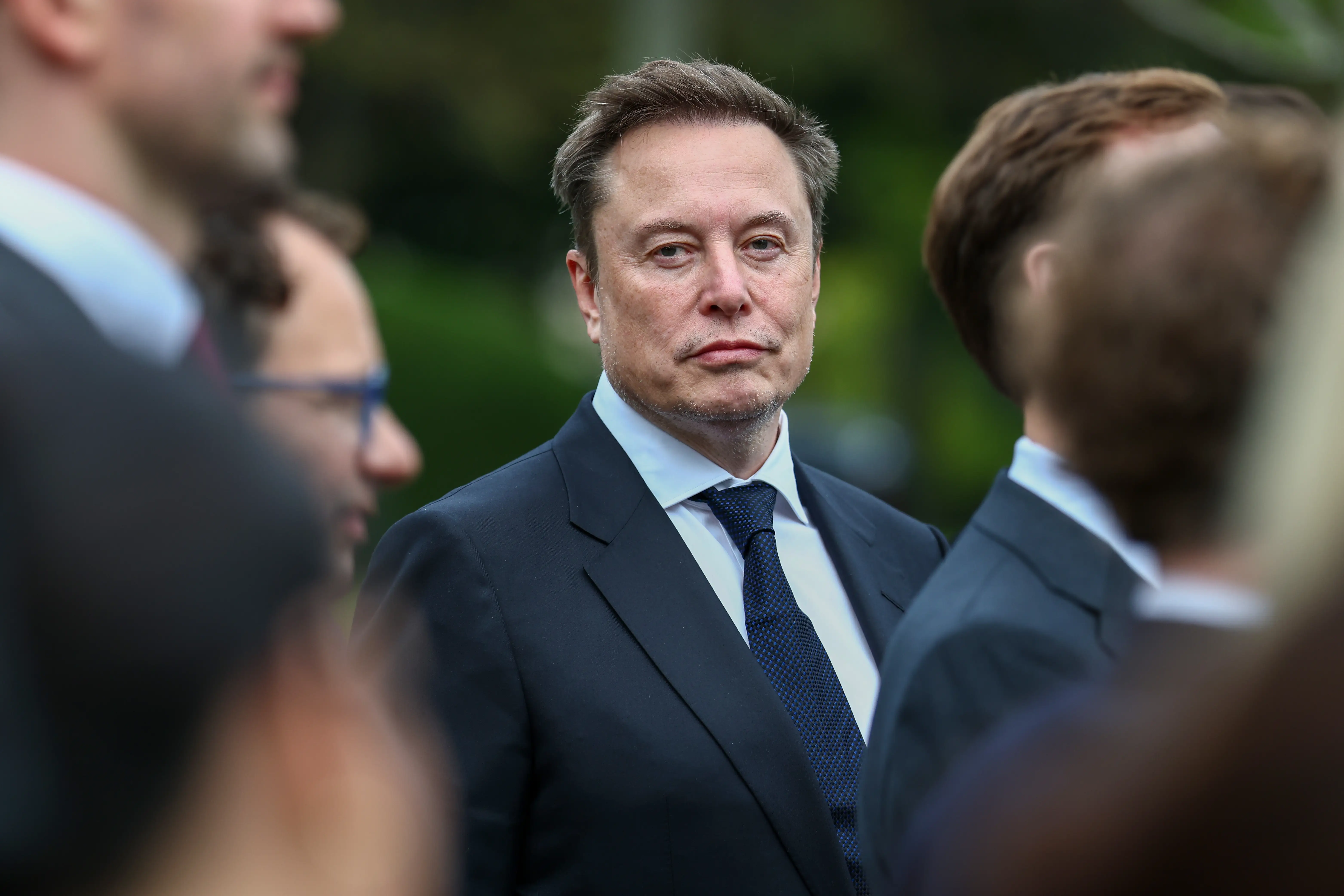 Elon Musk stands in a suit and tie amid a blurred outdoor crowd.