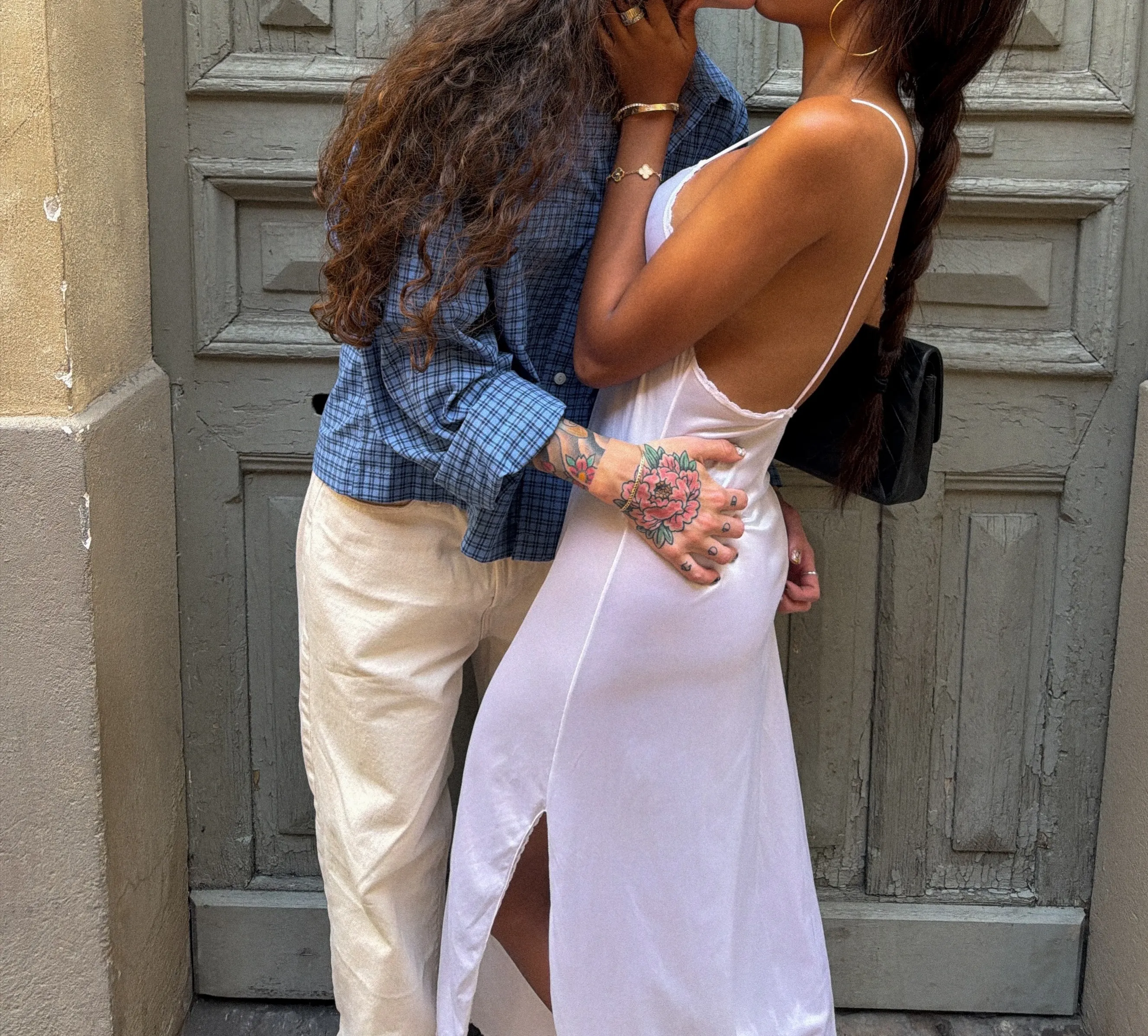 A couple kissing in front of a rustic wooden door, with one person wearing a blue plaid shirt and white trousers, and the other in a white slip dress.