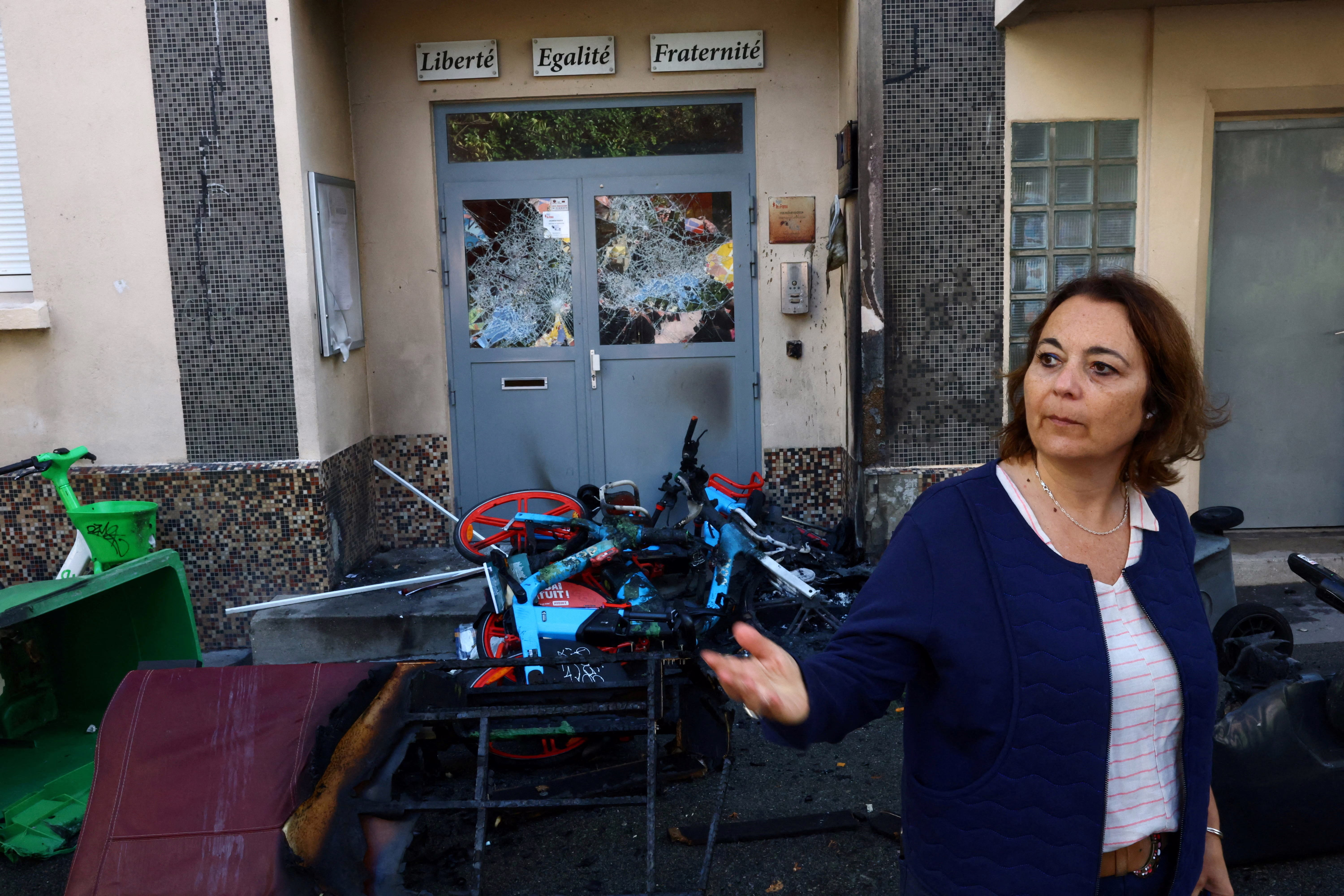 A high-school principal stands outside a school building beside damaged doors and debris, after a protest.