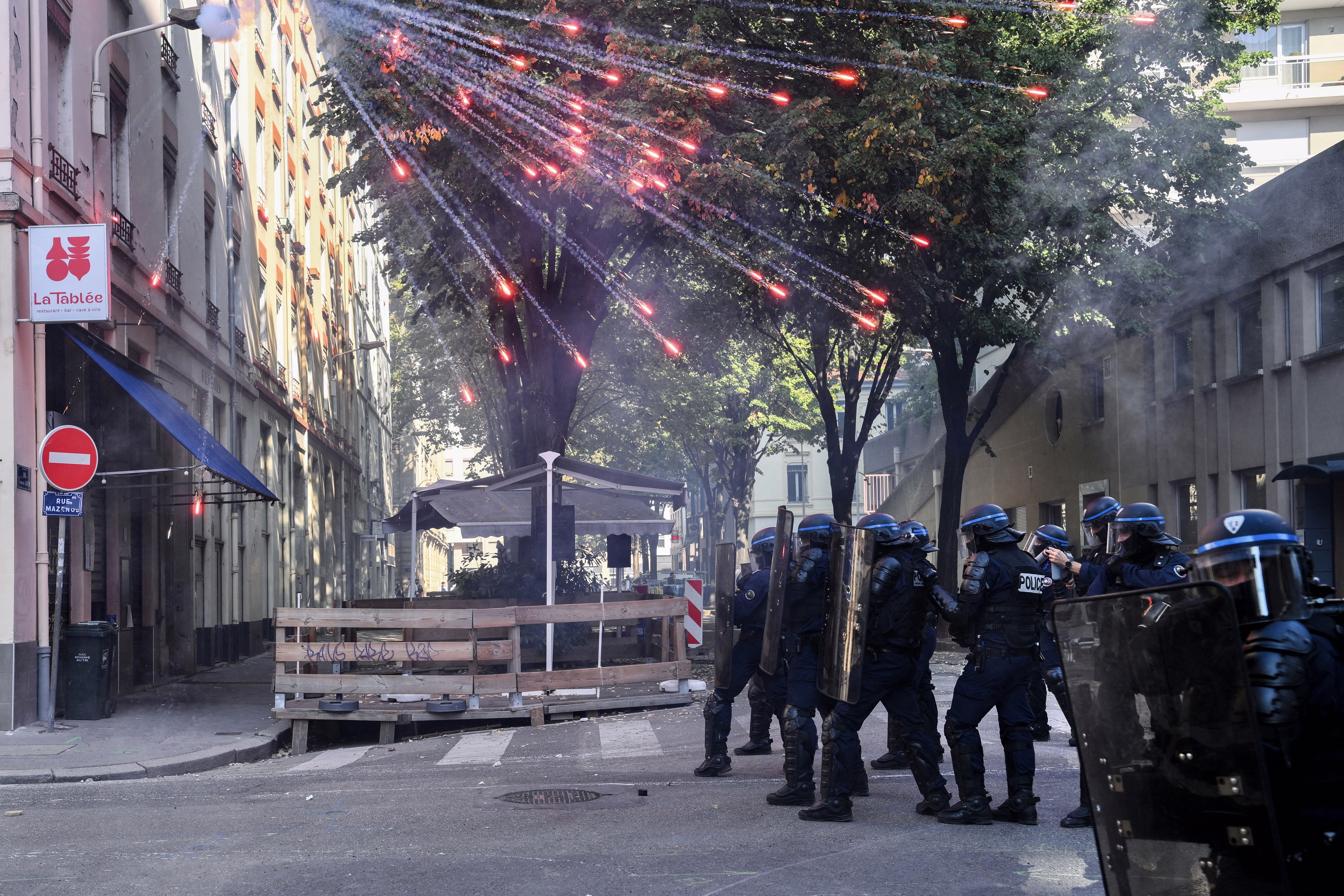 Riot police officers stand in a street as fireworks go off above them.