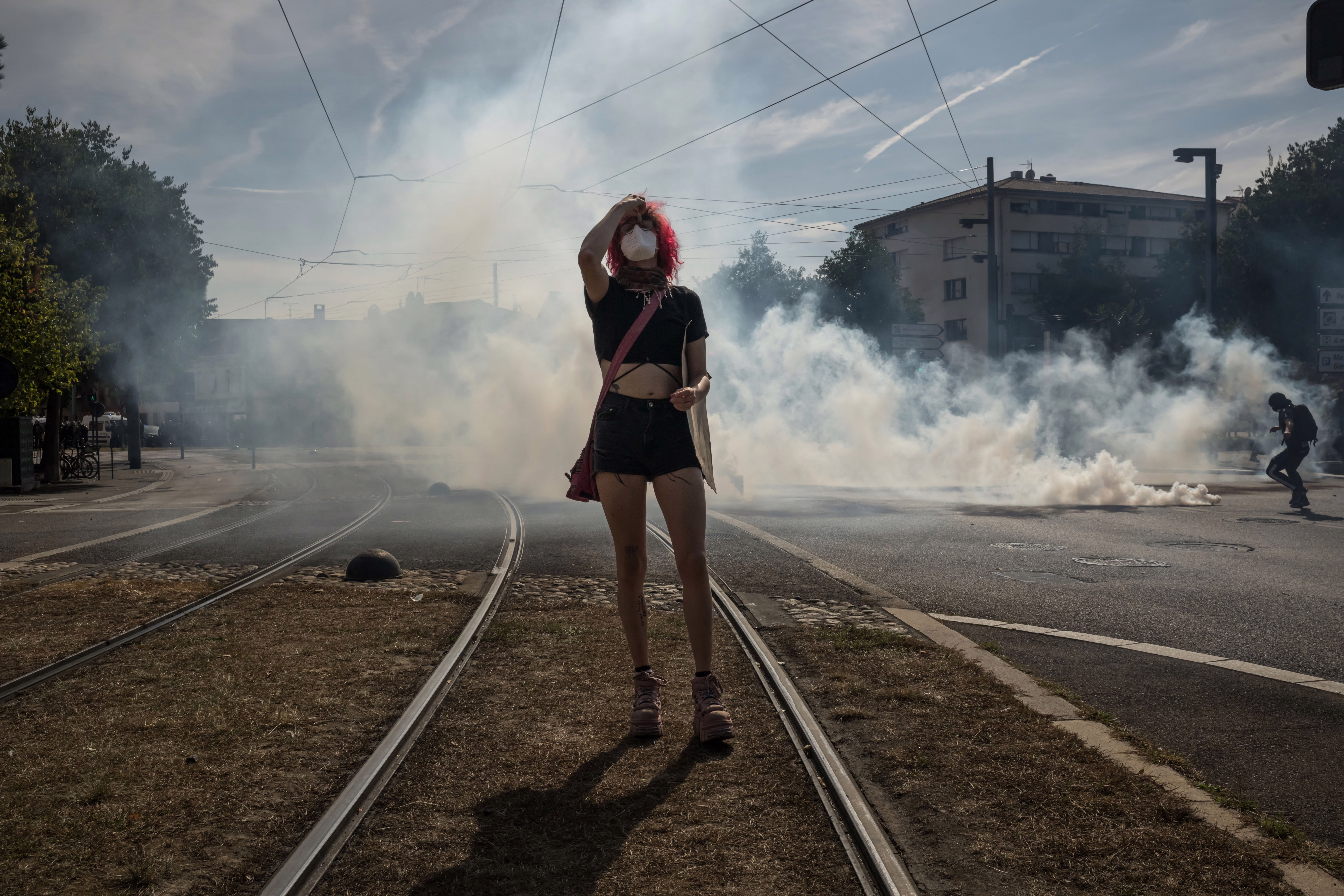 A student protester wearing a face mask walks, holding their head; cloud of tear gas is in the background.