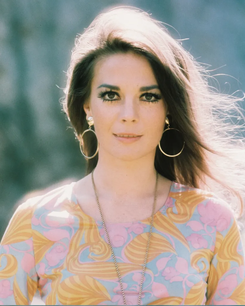 US actress Natalie Wood wearing a print pattern top with a large gold necklace and gold hoop earrings.