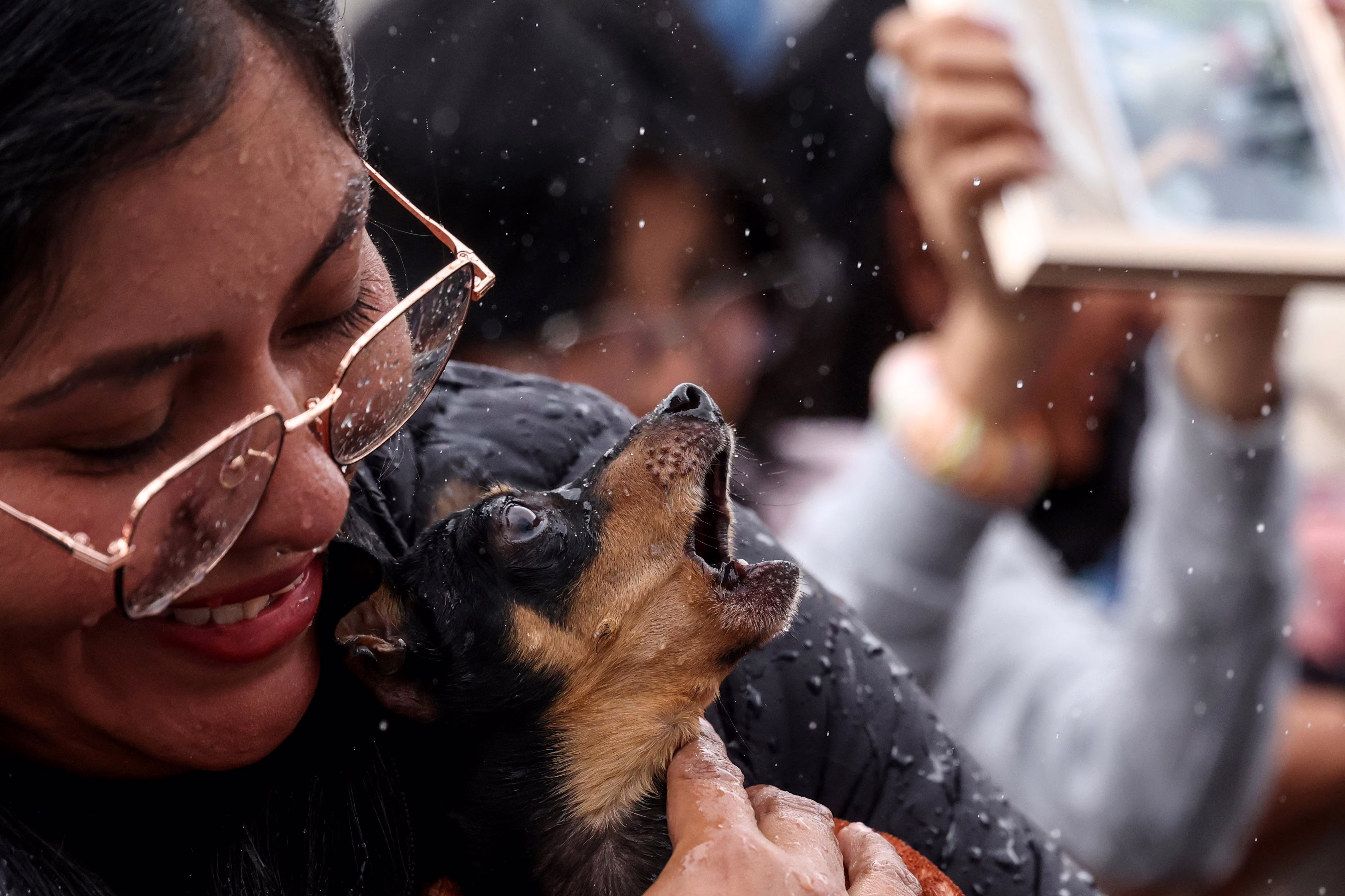 A woman holds a small dog that barks as it is sprinkled with holy water.