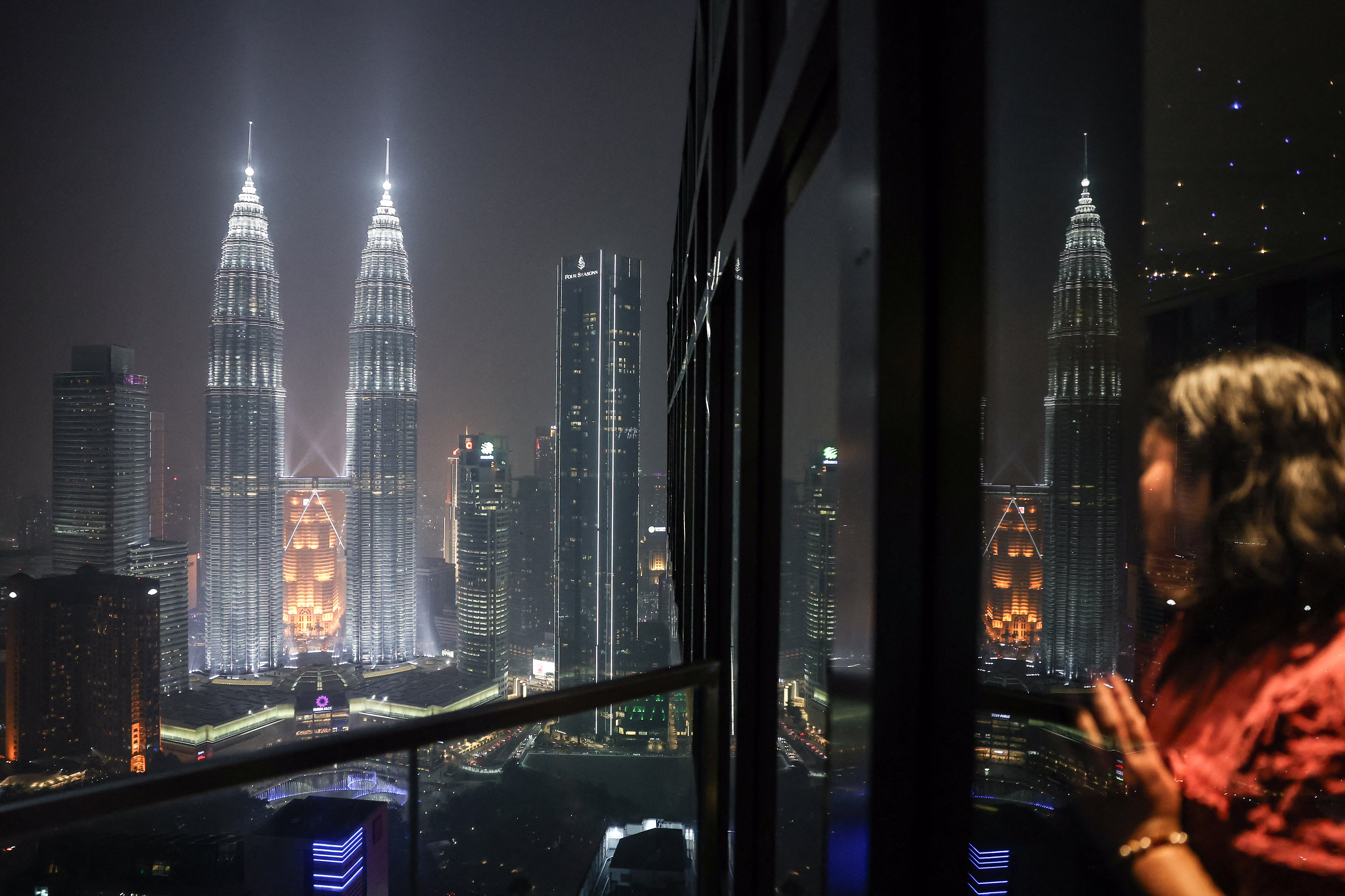 A woman looks out at the illuminated Petronas Twin Towers from a high-rise building.