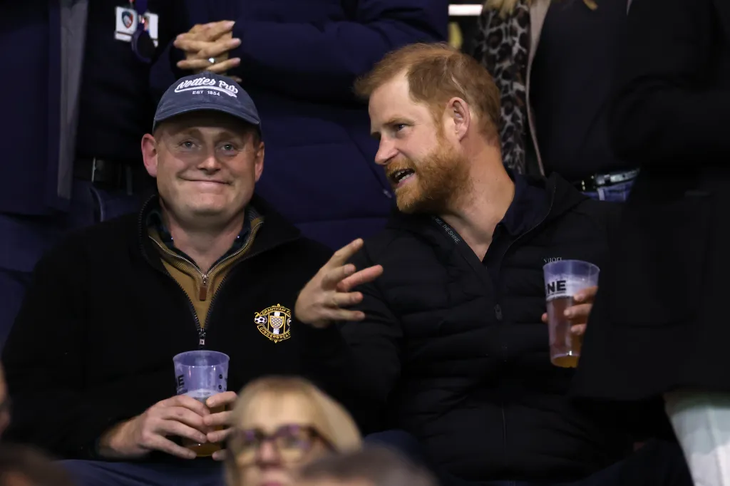 Prince Harry, Duke of Sussex sits in the stands and talks with another man while holding a drink during a rugby match.