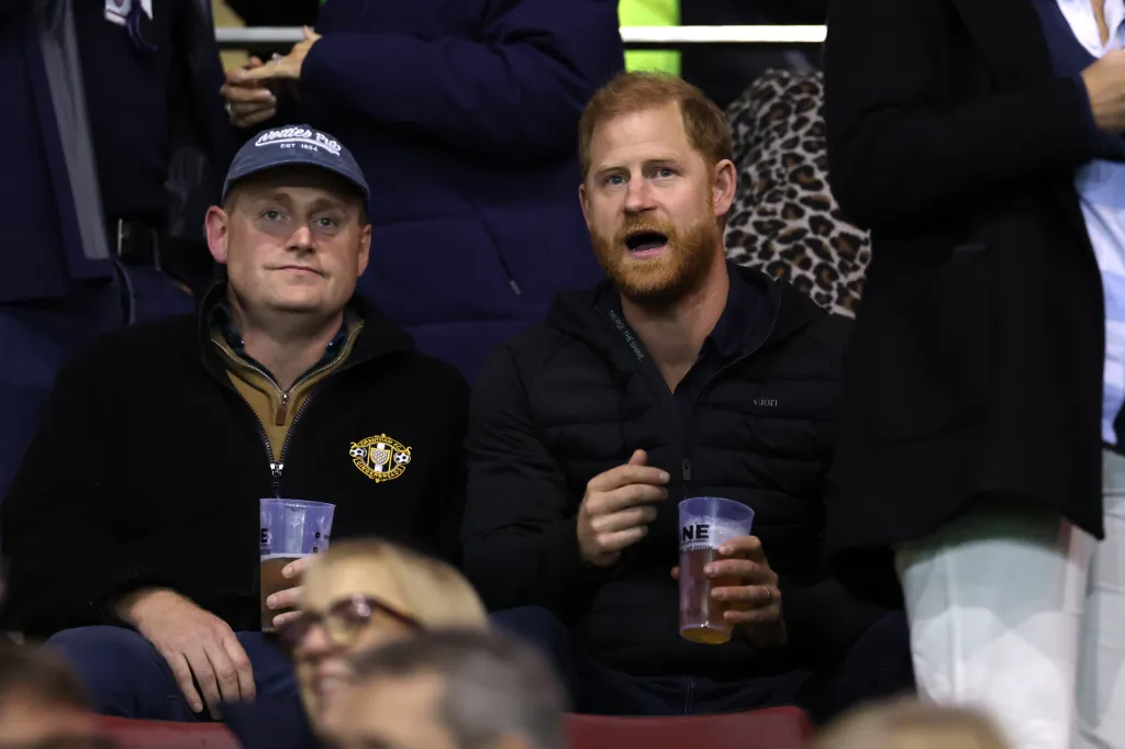 Prince Harry, Duke of Sussex sits in the stands at a rugby match, holding a plastic cup of beer.