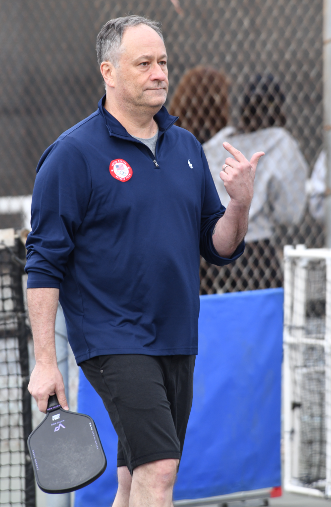 Doug Emhoff holds a pickleball paddle in a park.