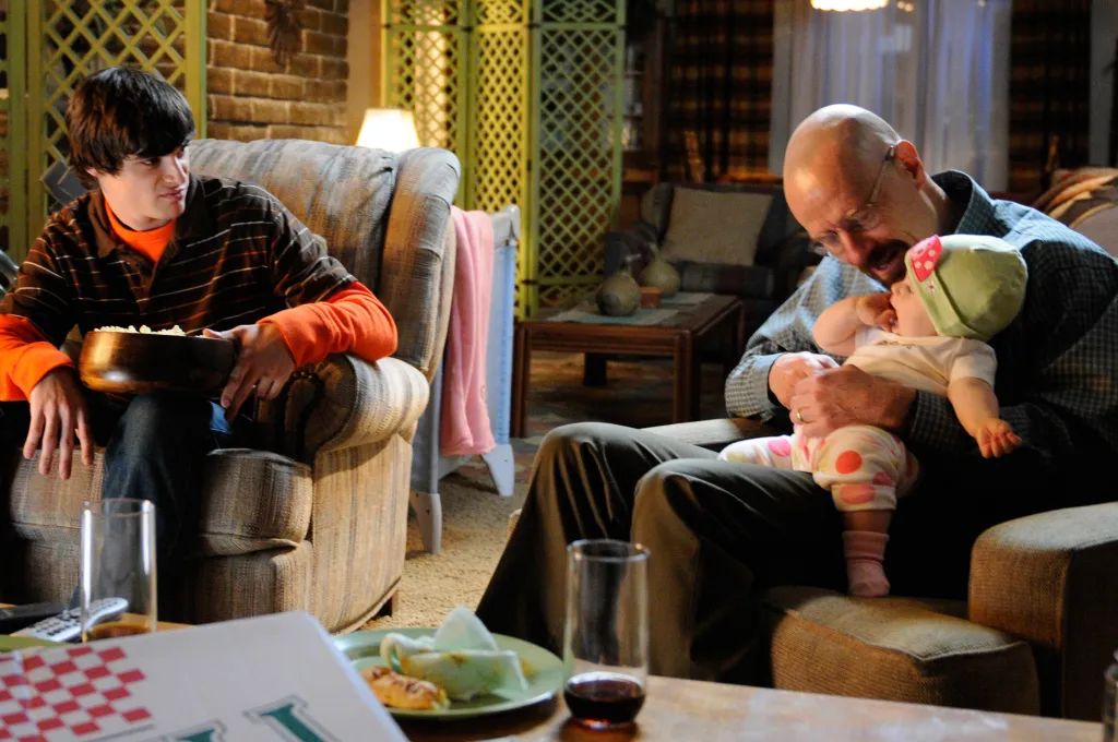 Bryan Cranston holding an infant while RJ Mitte sits nearby holding popcorn in a living room.