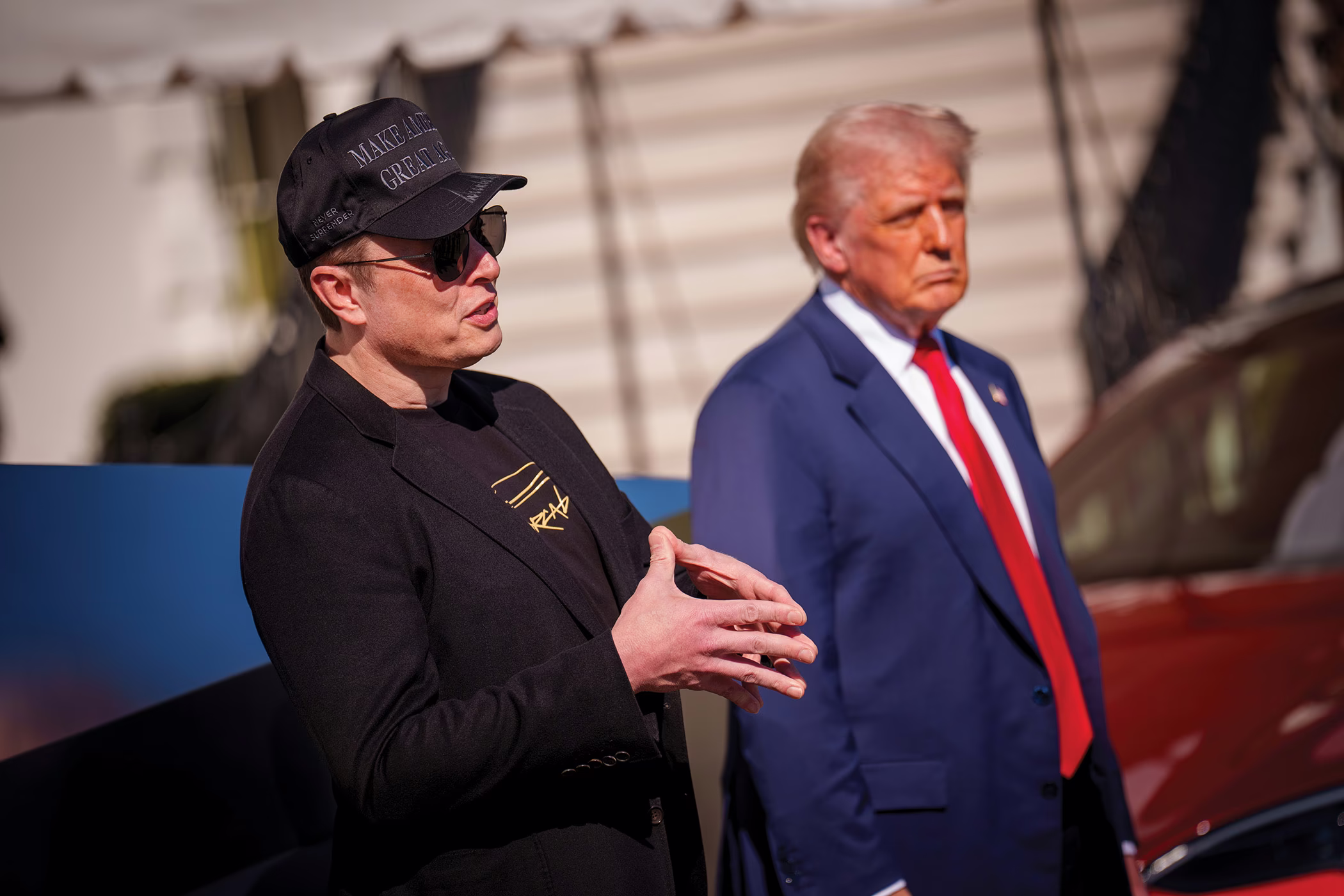 photo of Elon Musk wearing all-black 'Make America Great Again' baseball cap and sunglasses speaking at event next to Donald Trump in suit