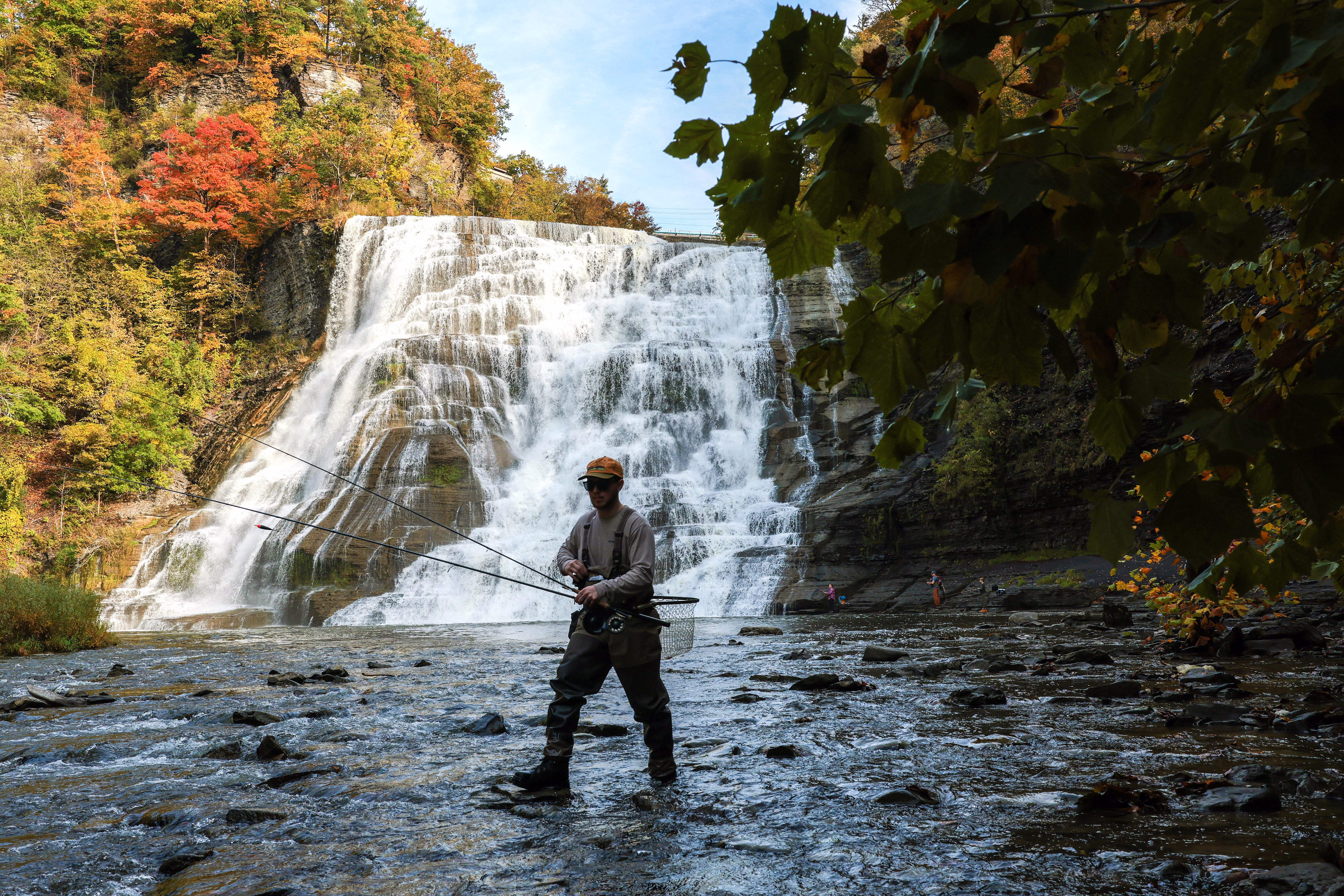 A person wearing waders fishes at the base of a broad waterfall.