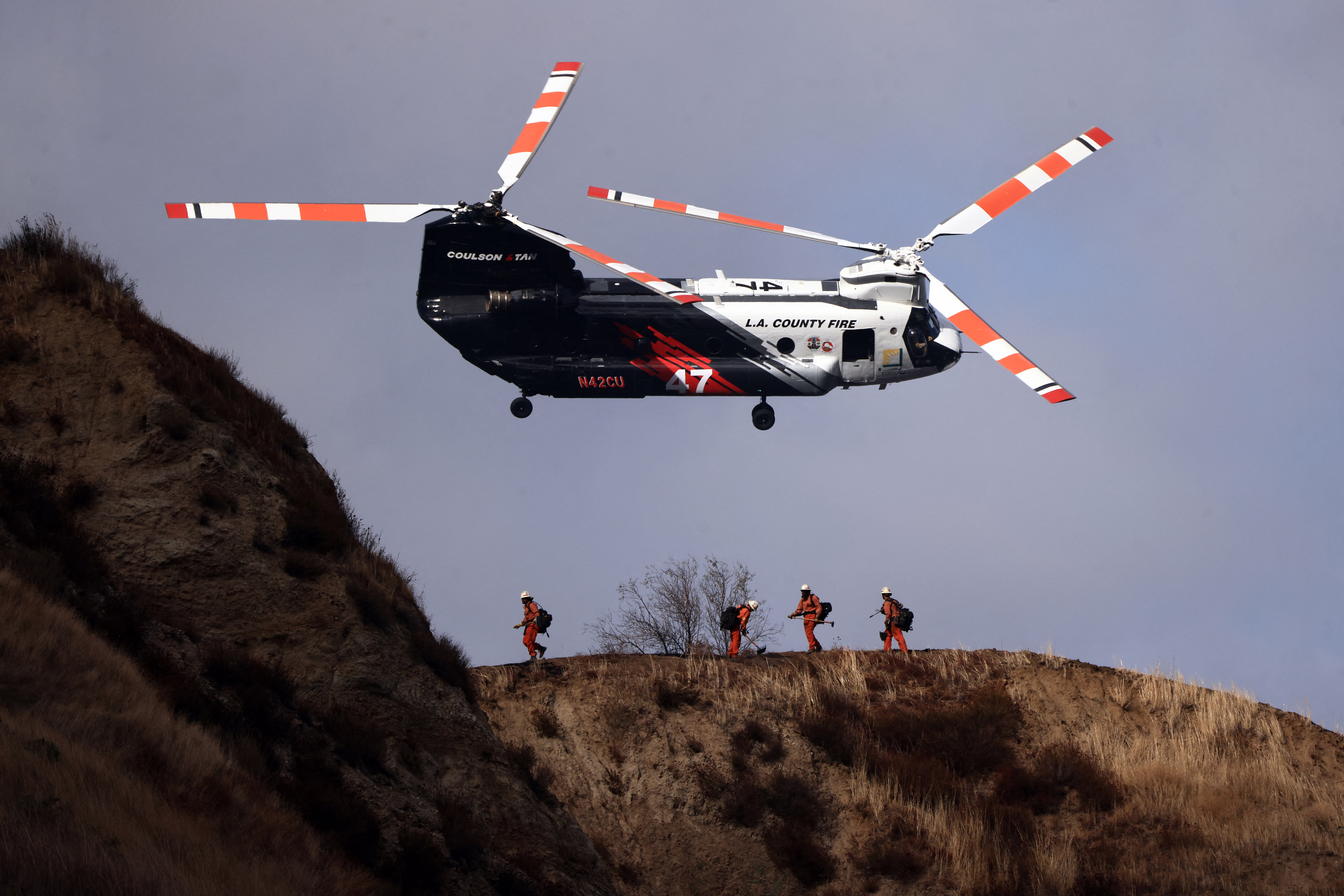 A firefighting helicopter flies above a fire crew on a hillside.