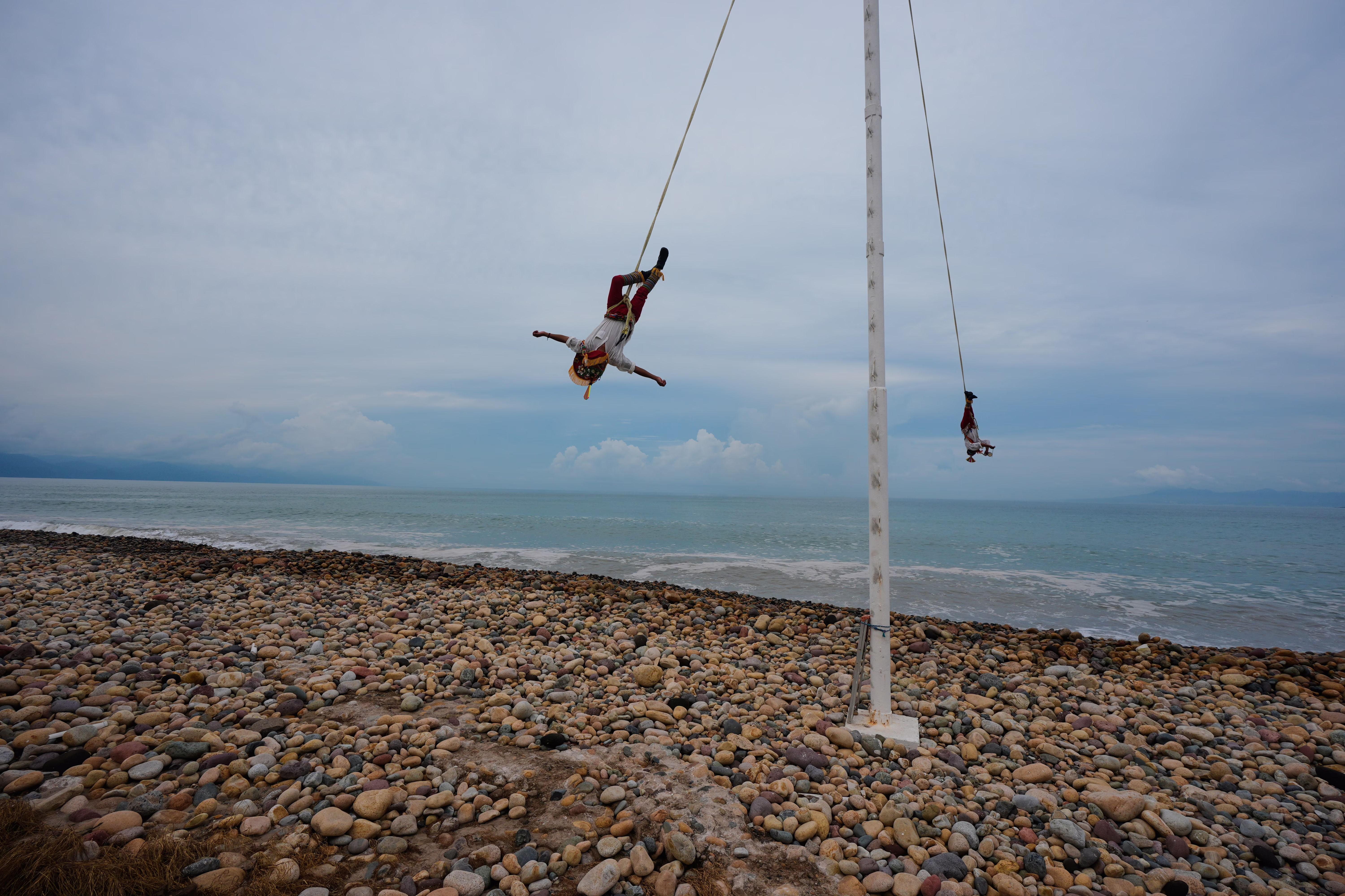 Two acrobats swing upside-down from ropes attached to a tall pool on a rocky beach.