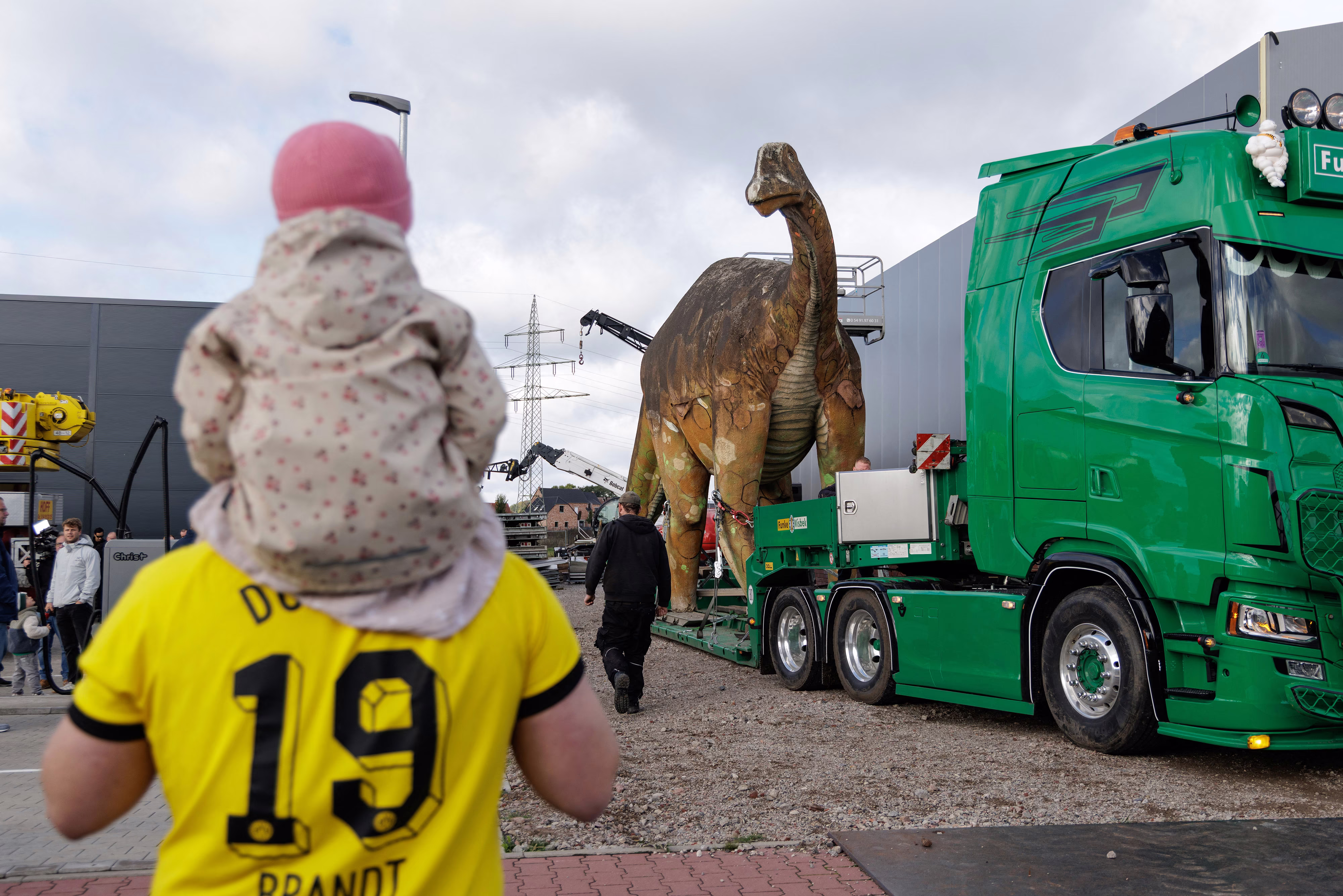 A life-sized dinosaur statue is loaded onto the low bed of a truck, as people watch.