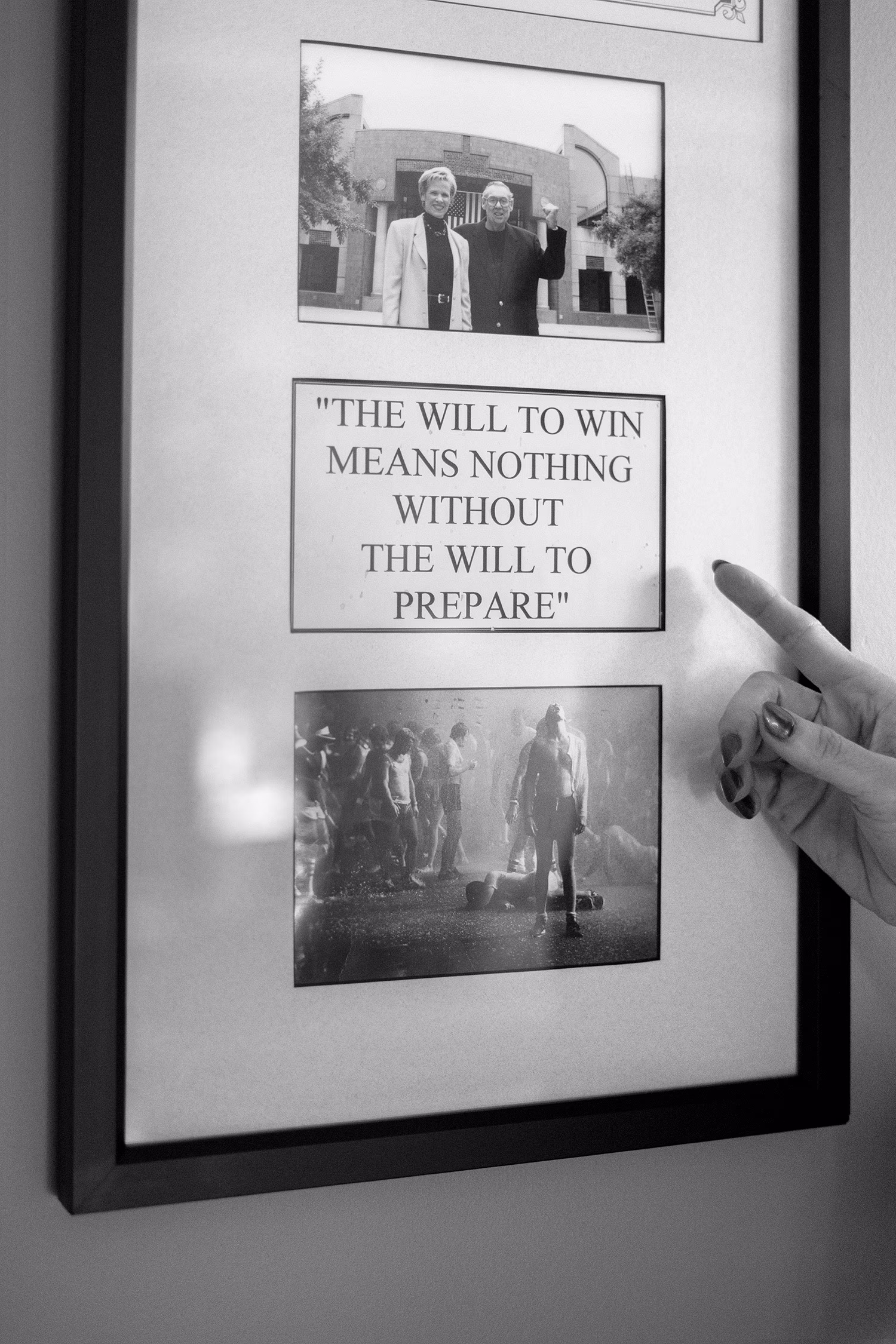A black and white photograph of Cyndy pointing to a frame photo with her mentor on the wall of her home. 