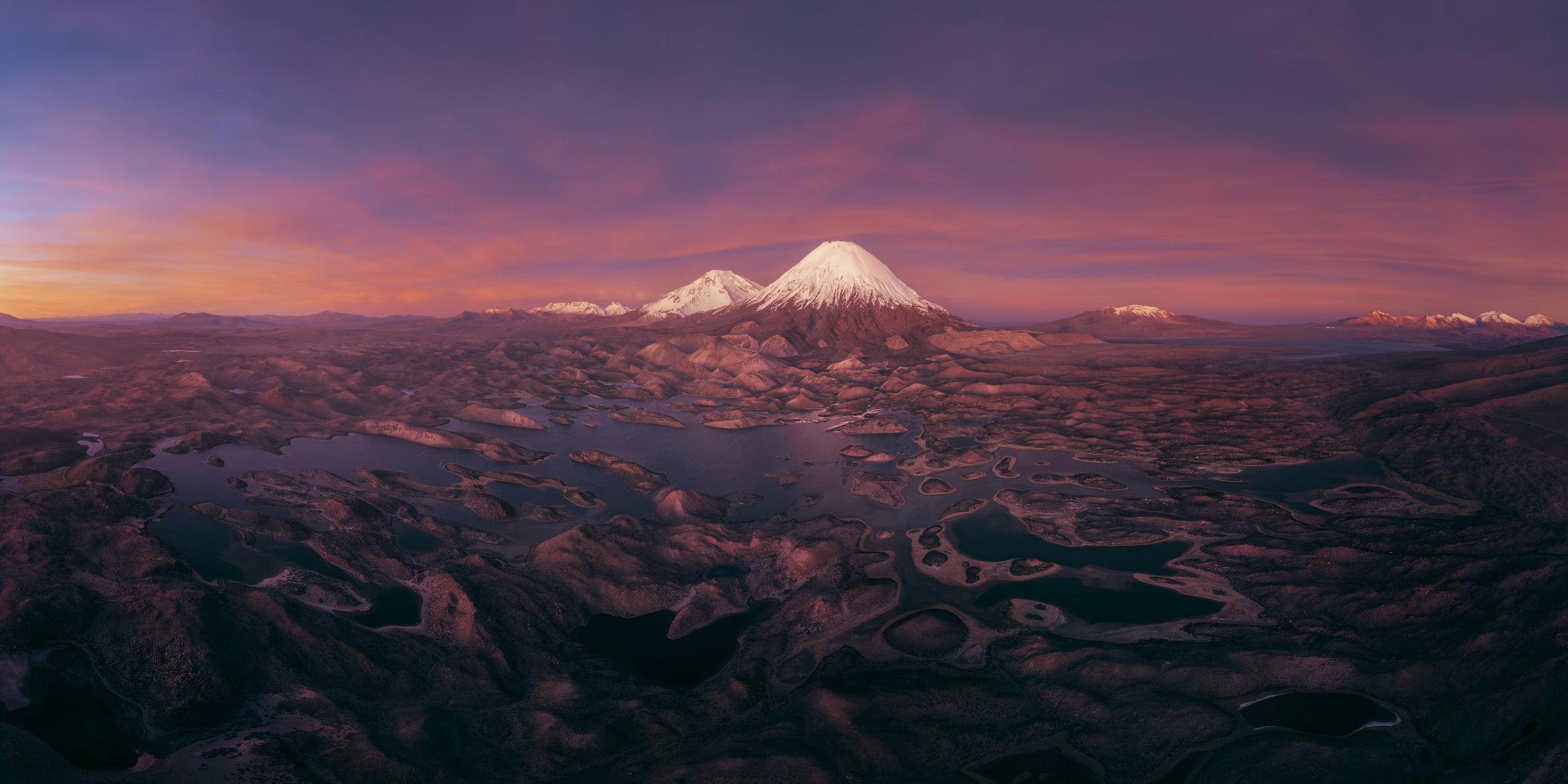 An aerial view of distant snowcapped mountains standing above an uneven field of foothills
