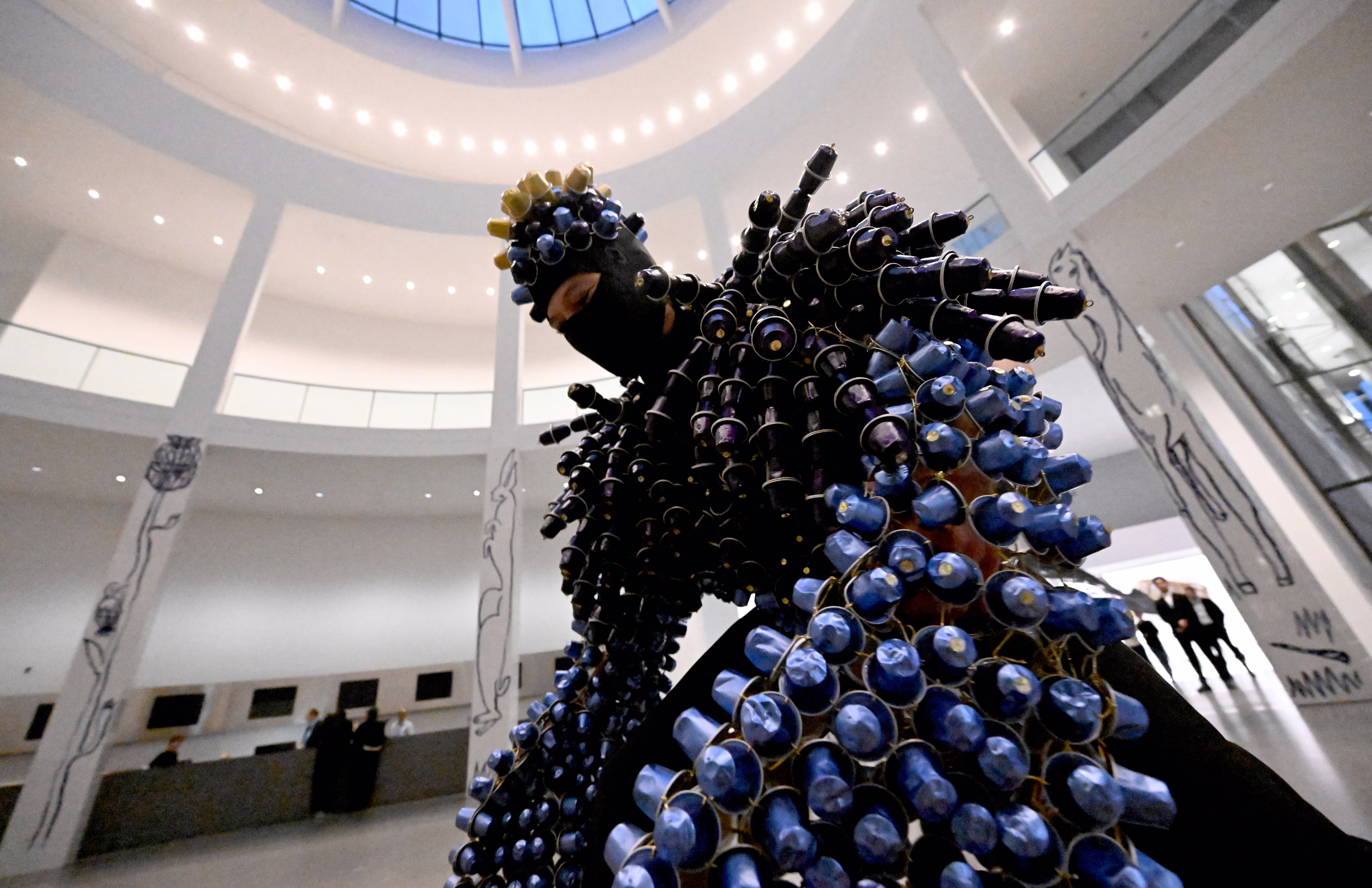A person wears a suit covered in what appear to be coffee pods during a performance in an atrium.
