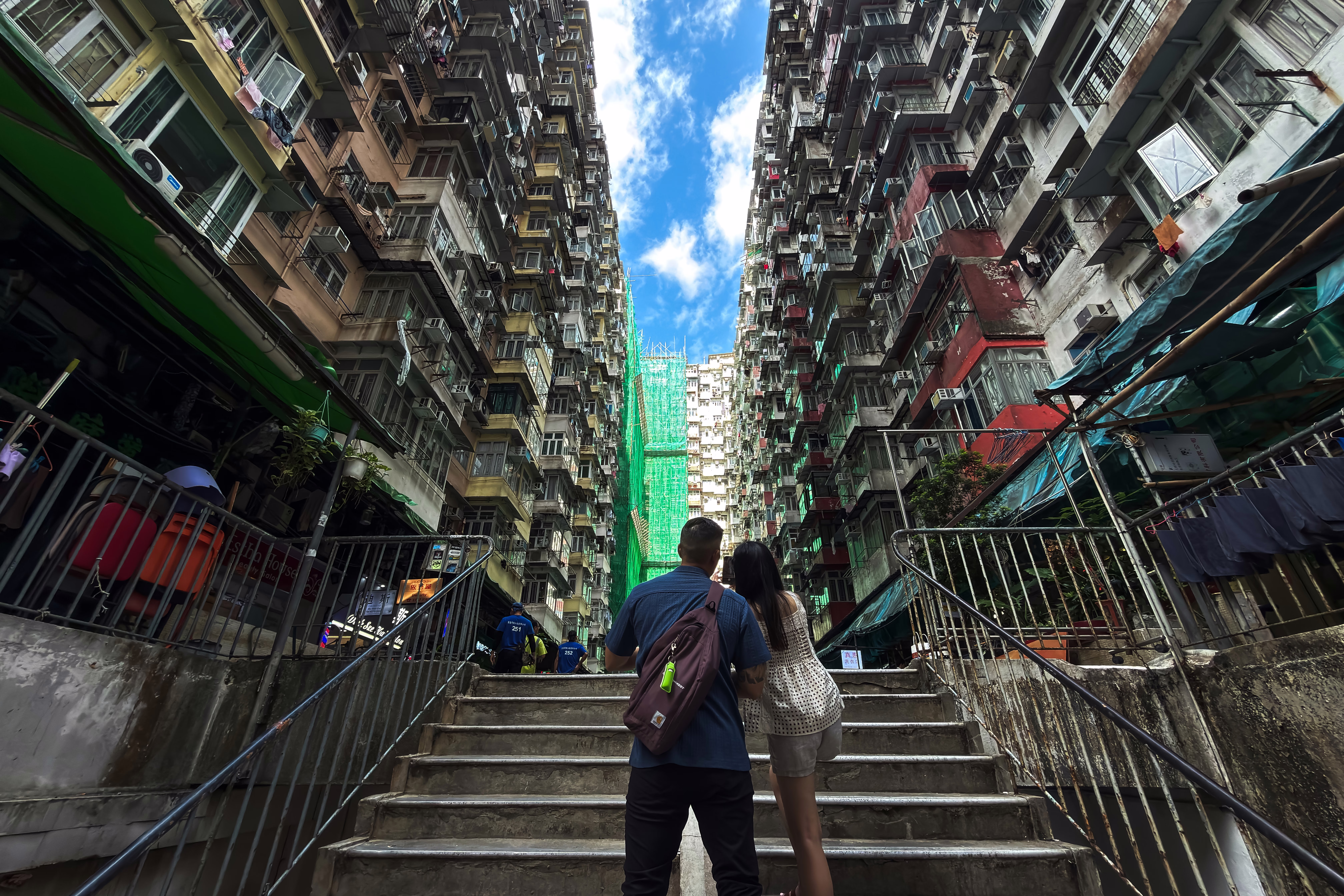 Two people stand on stairs, looking up at the sky between many tall, densely-built residential buildings.