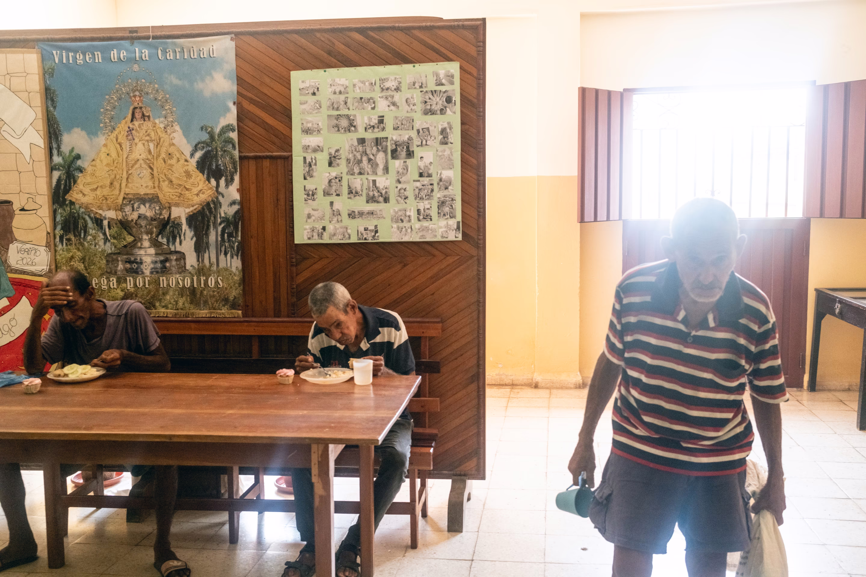 two elderly men sit and eat a meal while another man walks forward