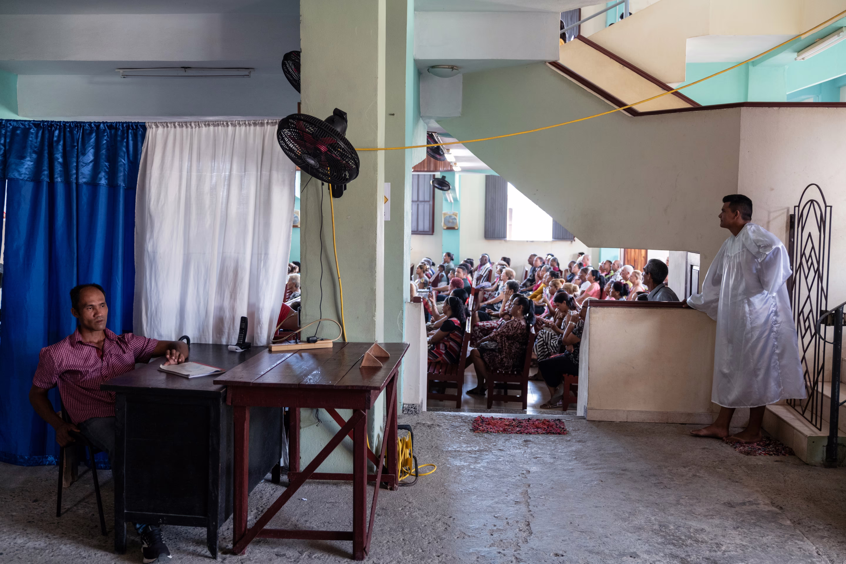 a man sits at a desk while dozens of people sit in seats behind him