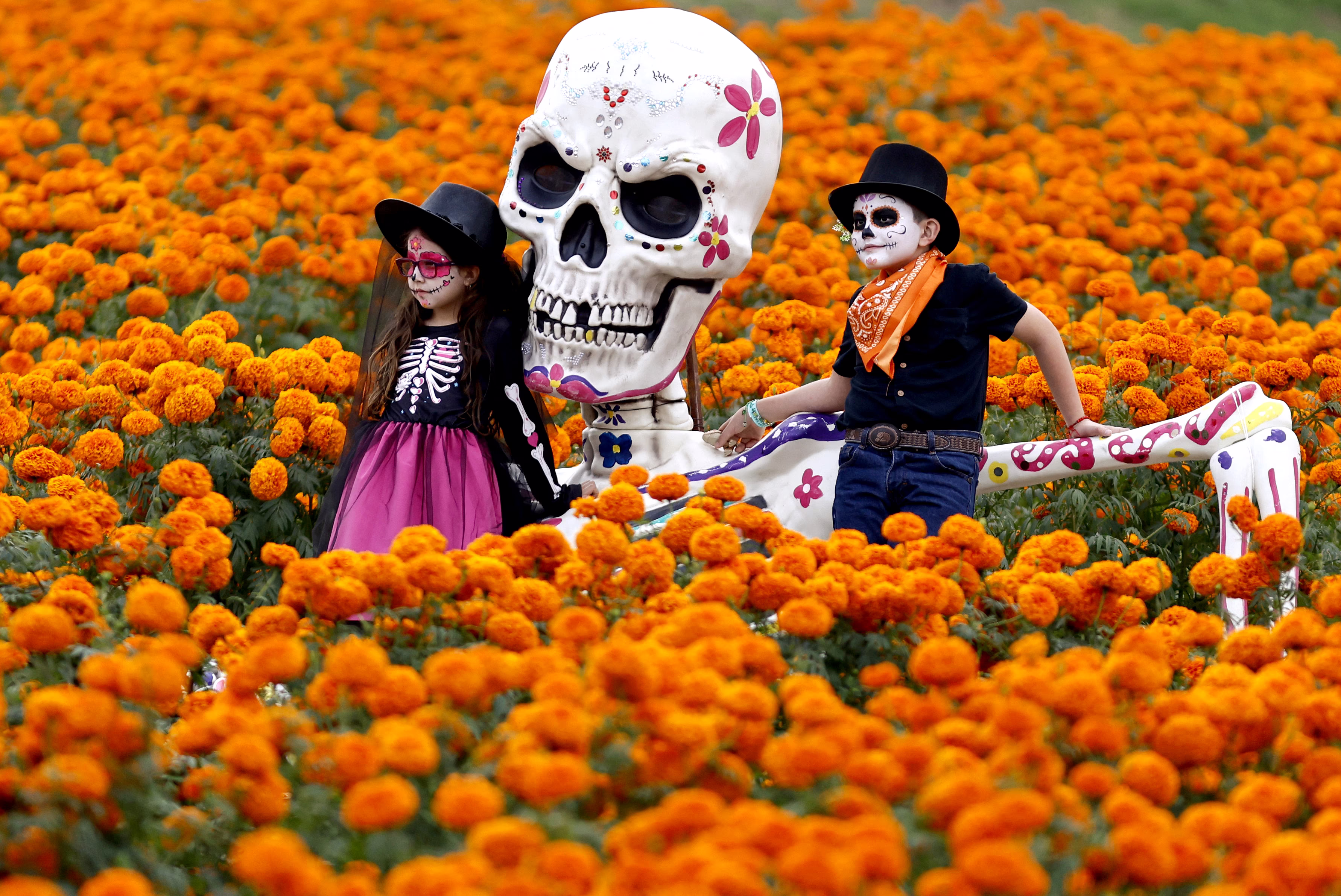 Two children with their faces painted pose for pictures in a field of flowers, beside a large skeleton statue.