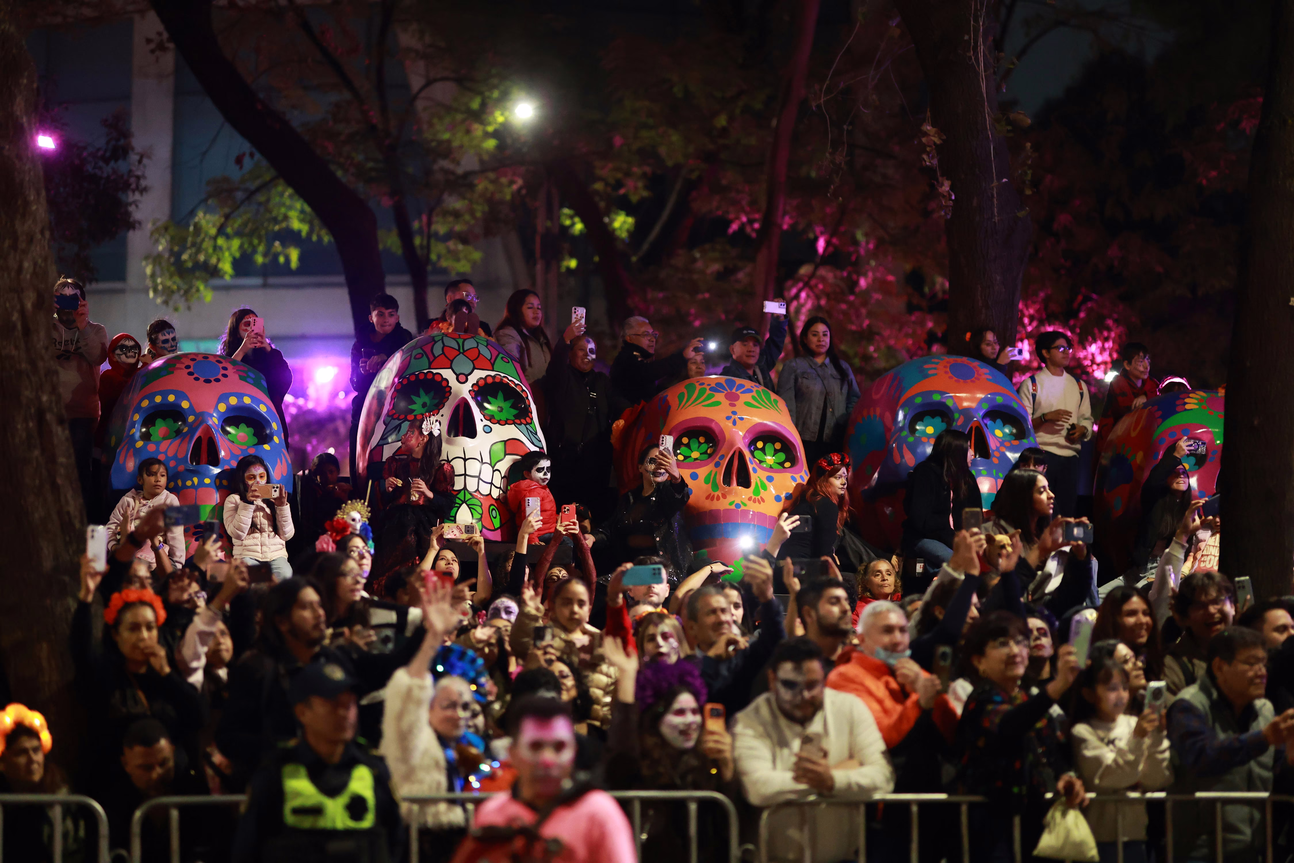 A crowd stands alongside several large skull sculptures, watching a passing parade.