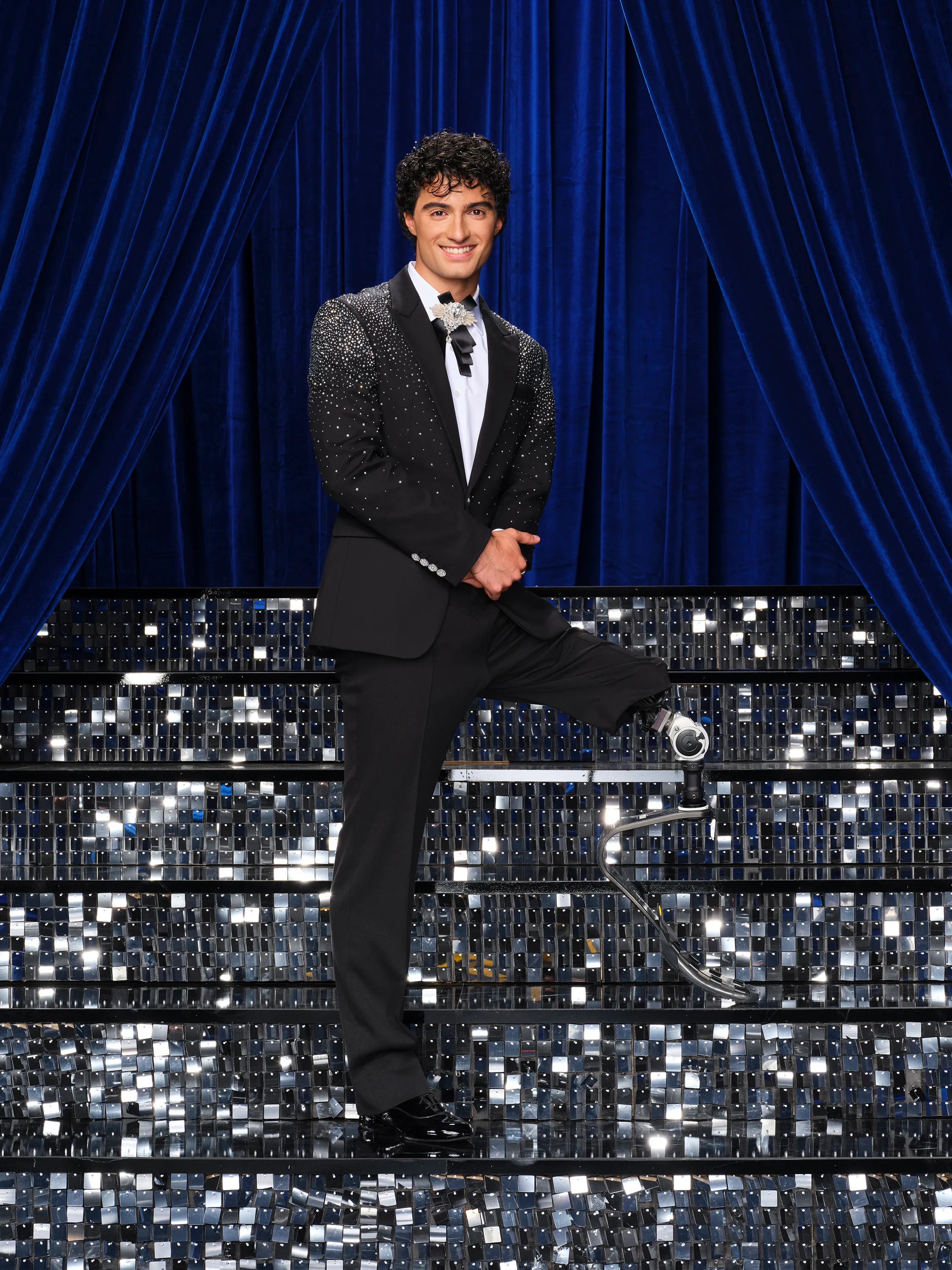 Ezra Frech in a black tuxedo with rhinestones on the shoulders, standing with one foot on a glittering staircase in front of a blue curtain.
