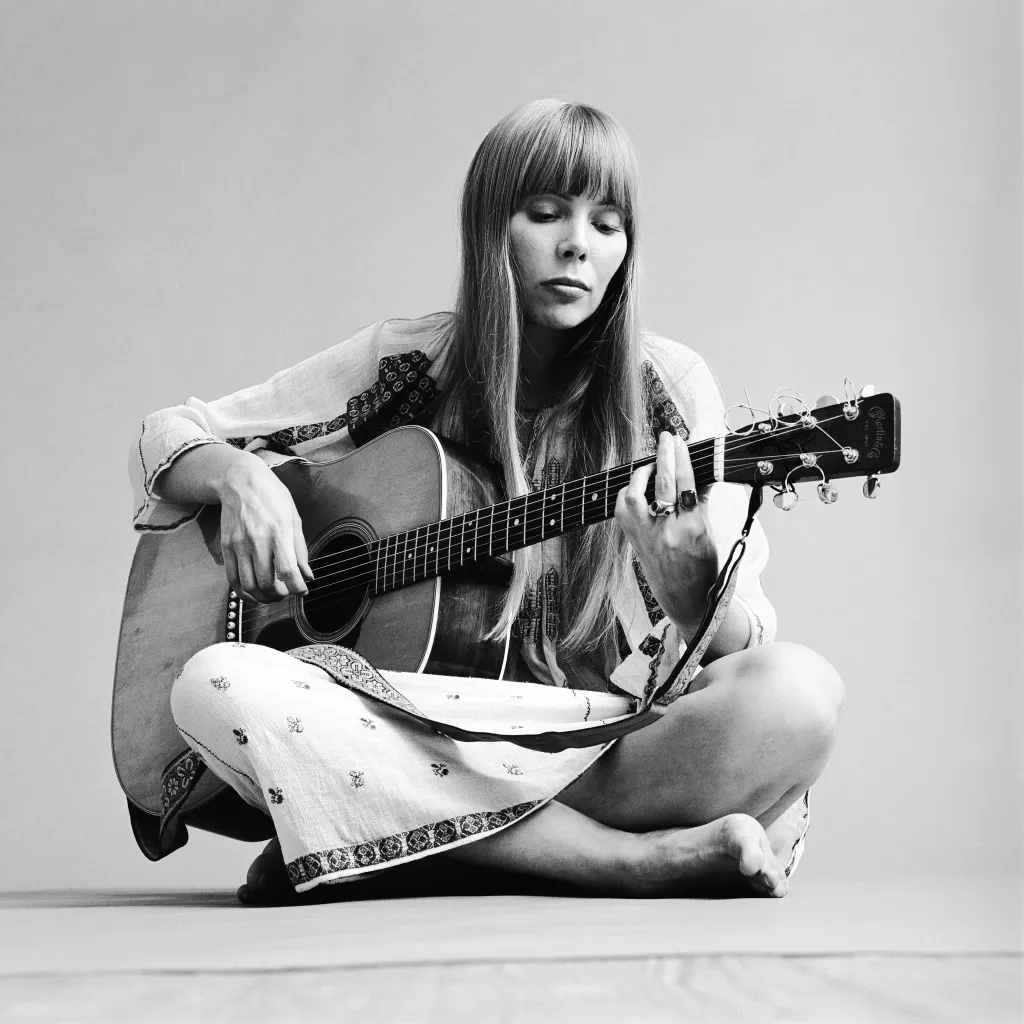 Black and white portrait of Canadian musician Joni Mitchell sitting cross-legged on the floor, playing an acoustic guitar.
