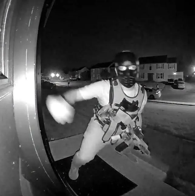 A doorbell camera view of a person wearing a mask and a tactical vest while carrying a rifle.