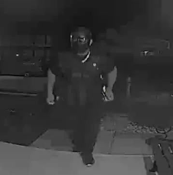 A grainy, low-light security camera image of a person wearing a mask and tactical vest approaching a home.