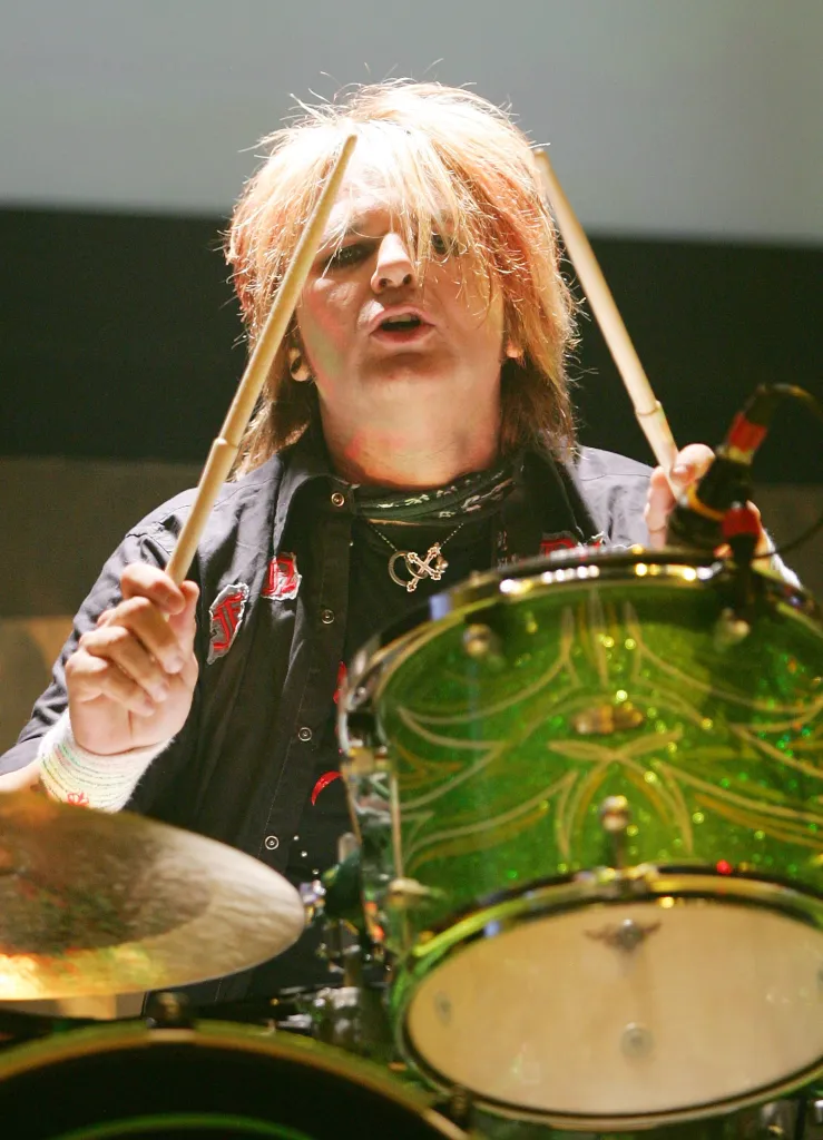 Poison drummer Rikki Rockett performs on stage, holding drumsticks above a drum kit.