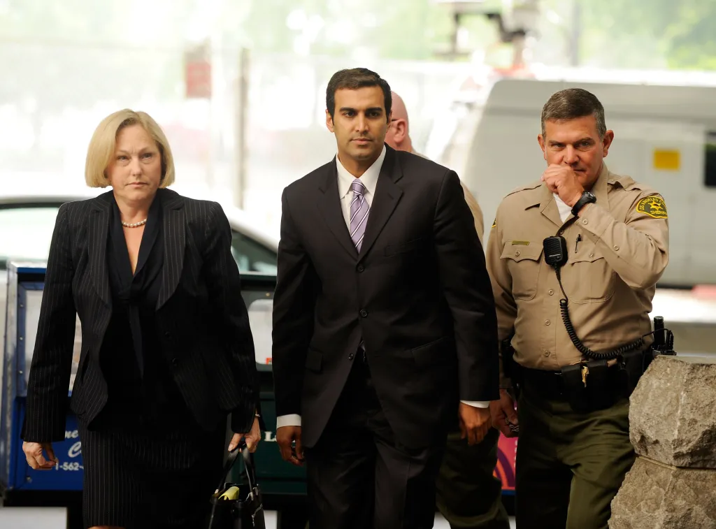 Dr. Sandeep Kapoo, in a black suit, arrives for his arraignment accompanied by Ellyn Garofalo in a black suit, and a law enforcement officer.
