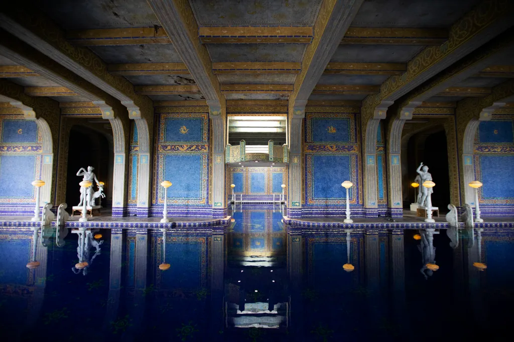 The Roman Pool at Hearst Castle, featuring blue and gold tiles, statues, and decorative pillars reflected in the indoor pool's water.
