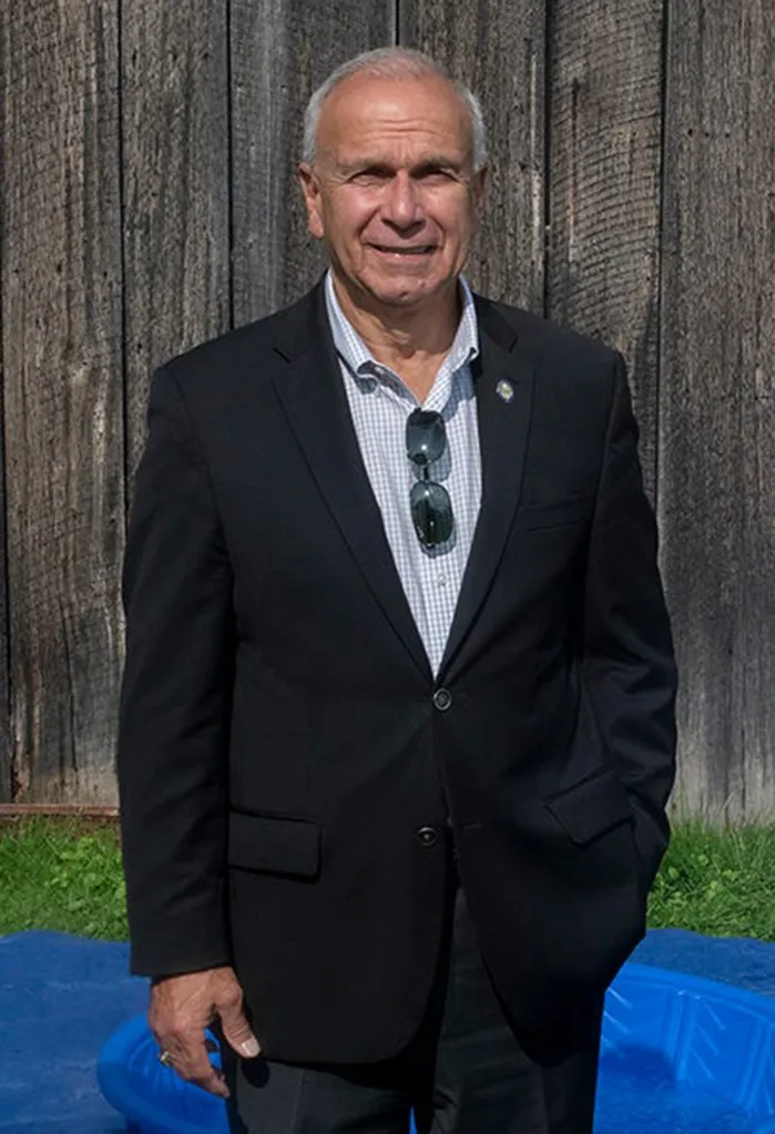 An older man in a black blazer and patterned shirt stands outdoors in front of a wooden wall.