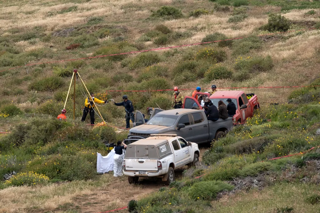 Rescue workers and forensics specialists gather near a waterhole and parked vehicles in a remote, grassy area where human remains were discovered.